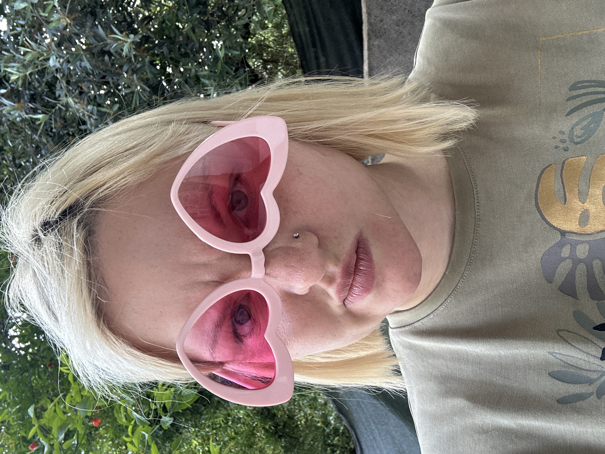 A woman with blonde hair and pink heart-shaped sunglasses is angled toward the camera. She has a nose piercing and is wearing an olive green t-shirt with a gold and gray design. The background consists of foliage, possibly trees or bushes. This image appears to be taken outdoors. The location in the city of Budva, Montenegro is not discernible from the image.