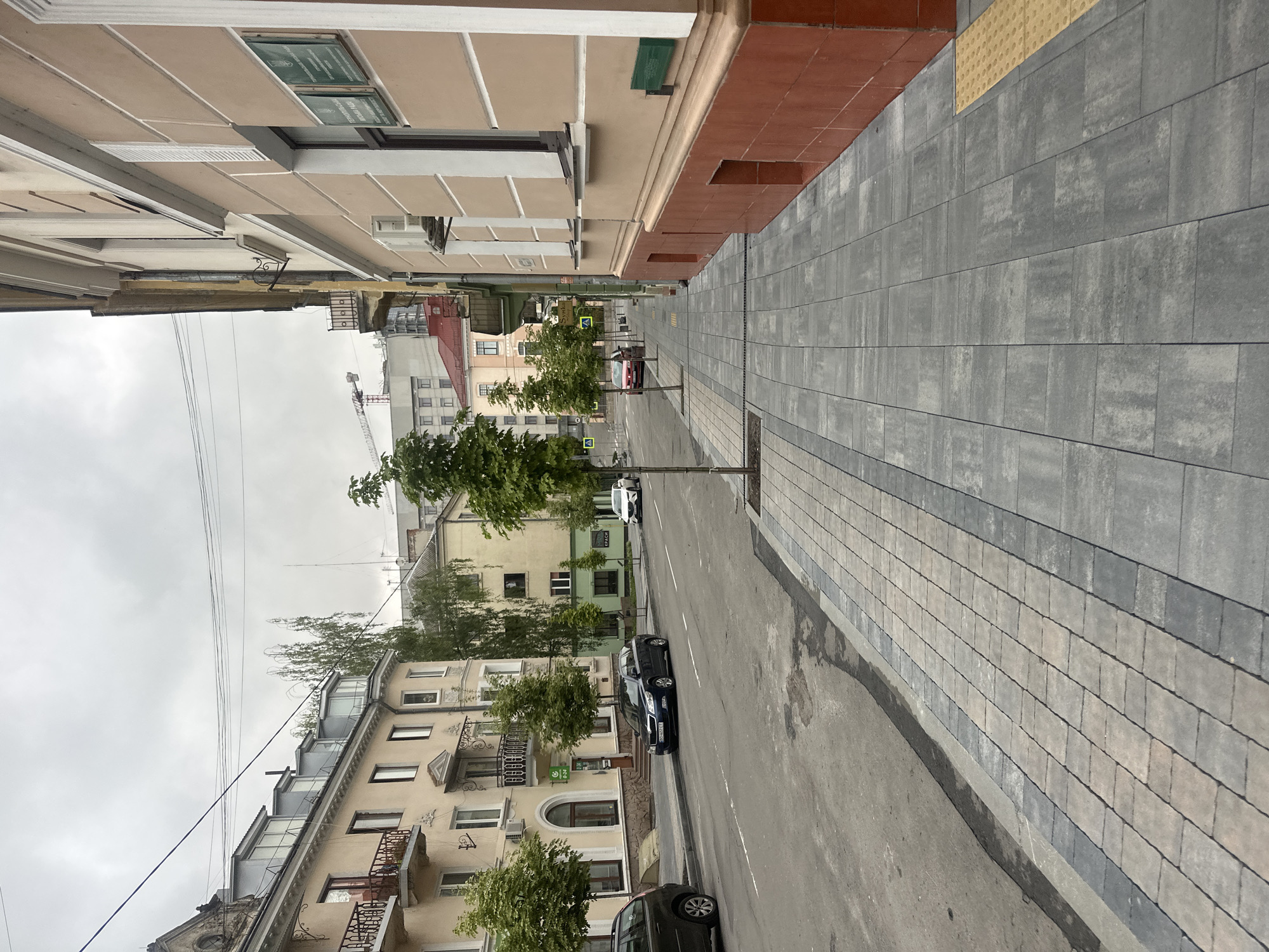 The image captures a street view in Ternopil, Ukraine, on an overcast day. The perspective is from the sidewalk, looking down a street lined with buildings and trees. On the right, a light beige building with a red-brown foundation dominates the foreground, featuring windows and some air conditioning units. Signs are visible on its facade. To the left, several buildings of similar architectural style, painted in lighter colors, are present, with balconies and windows. The street itself is paved, with parked cars visible. Trees are planted along the sidewalk, providing shade. In the background, more buildings, some under construction with cranes, are visible. The sky is cloudy.
