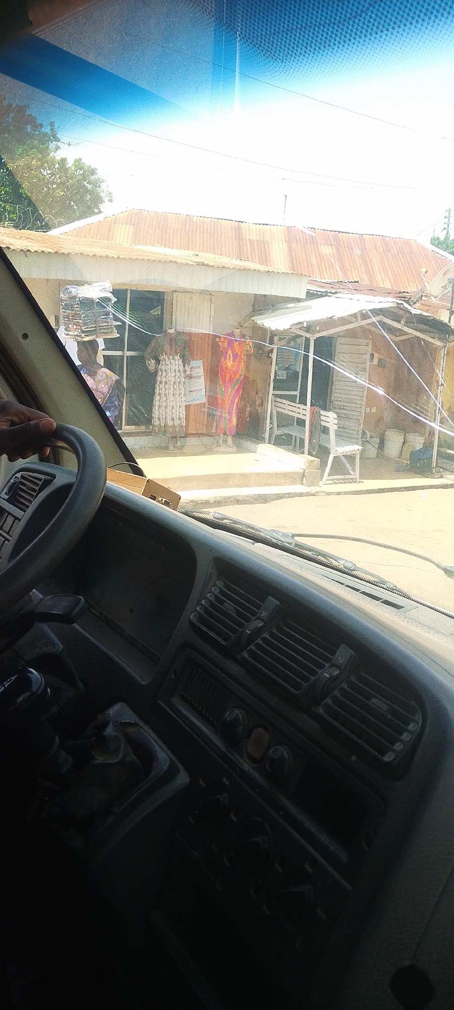 The image, taken from inside a vehicle in Jos, Nigeria, shows a street scene. In the foreground, a hand is visible on the steering wheel, and part of the vehicle's dashboard is visible, including air vents and controls. Outside the vehicle, a shop with a corrugated iron roof is prominent. Several items are displayed in front of the shop, including two mannequins dressed in patterned fabrics and a stack of folded textiles being carried on a person's head. In front of the shop, there is a covered outdoor stall with a wooden bench. Two white buckets are also visible near the stall. The overall environment suggests a commercial area in Jos, characterized by simple structures and street vendors.