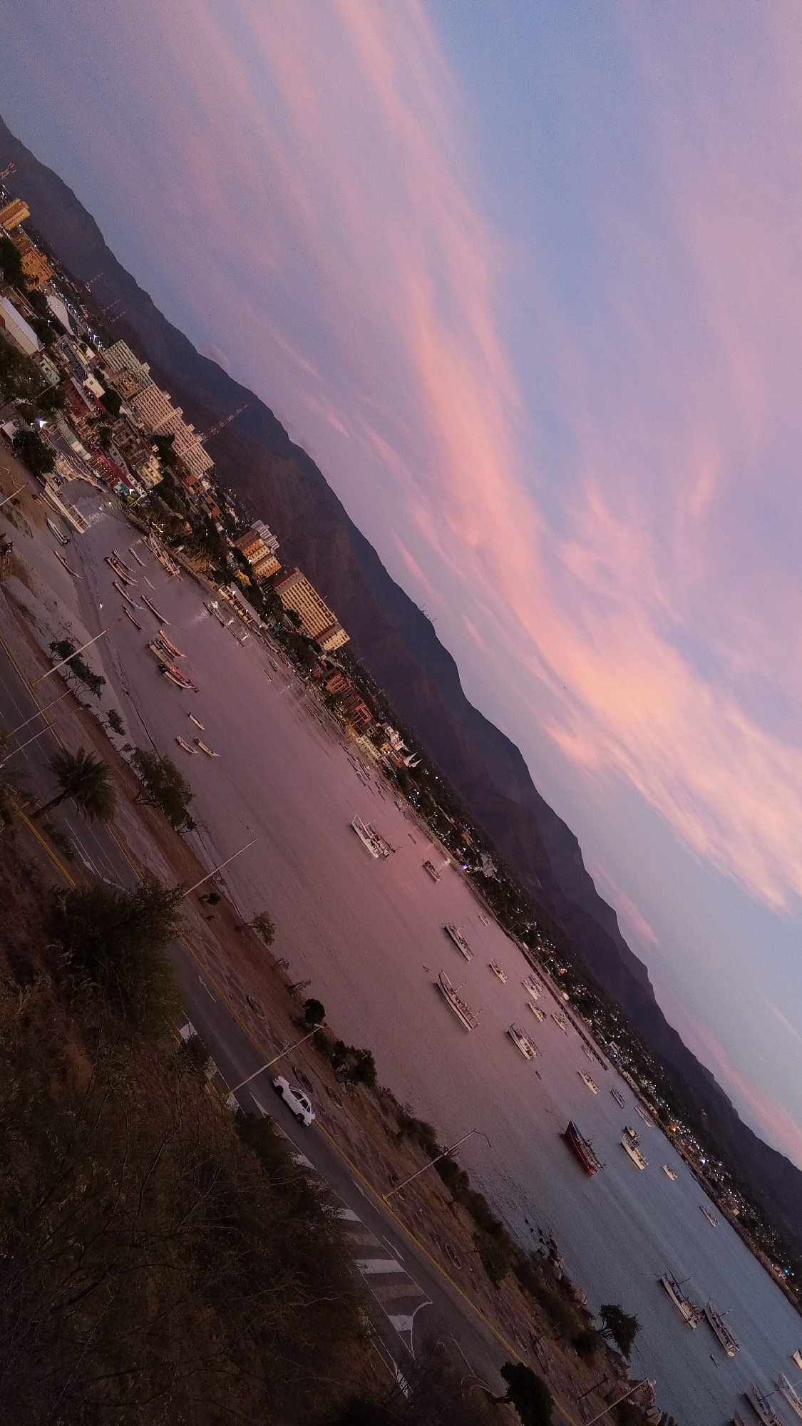 The image shows a view of Juan Griego, Venezuela, at sunset. The camera is angled, creating a diagonal composition. In the foreground, a road runs alongside the water, with a white car visible. The water is filled with numerous boats and small vessels. The city buildings are concentrated along the shoreline, with taller structures mixed in. The backdrop features a mountain range. The sky is colored with pink and purple hues, indicating the time of day. The overall environment is serene.