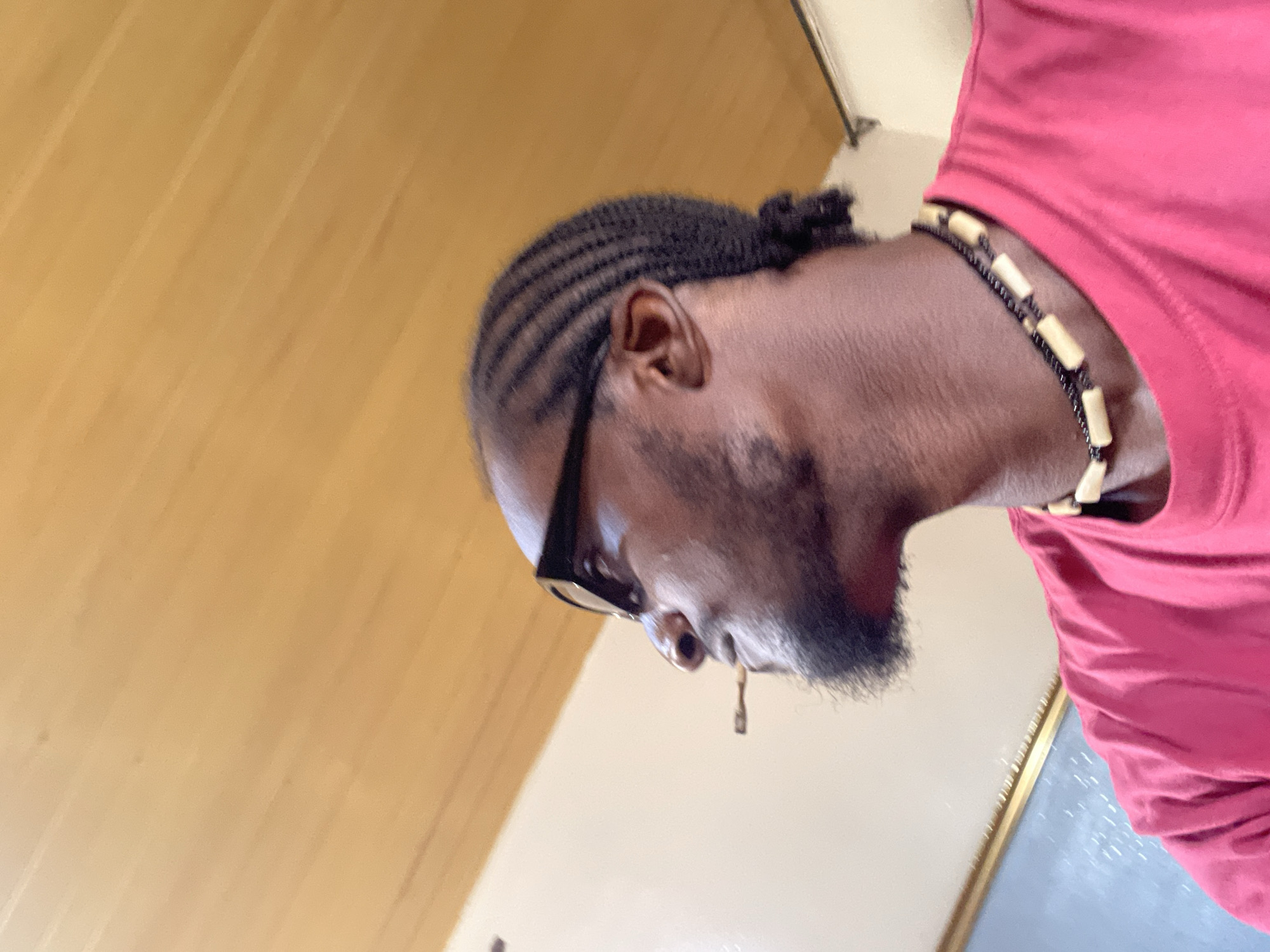 A medium close-up, low-angle profile shot depicts an adult male of African descent, positioned with his head tilted slightly upwards and to the left. He is wearing dark-framed eyeglasses and has dark hair styled in cornrows pulled back into a bun. A dark goatee is present on his chin. A light-colored, cylindrical object, consistent with a cigarette or cigarillo, is held between his lips on the left side of his mouth. He is adorned with a two-strand necklace featuring small dark beads and larger, rectangular, light-colored beads. He wears a plain red short-sleeved t-shirt. The background comprises a light brown wooden plank ceiling above a light-colored wall. A reflective surface with a blue tint is partially visible in the lower left, and a small white fixture is on the far right. The image does not contain specific visual identifiers to ascertain a precise location within the city of Jos, Nigeria.