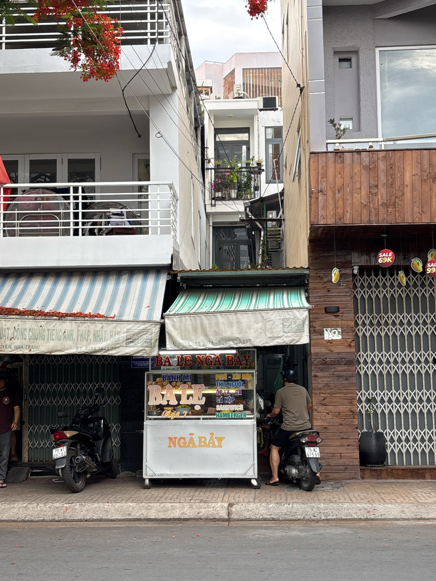 The image captures a street scene in Nha Trang, Vietnam, with a focus on a food stall and the surrounding buildings. The food stall, with a white metal cart, is illuminated and displays various food items and menu options including "Banh Mi", "Pate - Xiu Mai", "Thit Nguoi", "Op La Ga", "Ca Hop", "Sandwich", and "Hamburger". A man wearing a dark green t-shirt and black shorts, with a black helmet, is seated on a black scooter behind the stall, appearing to be a vendor or customer. Another scooter is parked to the left of the stall, partially obscured by the stall itself. In the background, there are multi-story buildings. The building to the left has a white facade with balconies featuring white railings and is adorned with red flowers hanging from above. The building to the right is constructed with wooden planks and has a large metal gate with a diamond pattern. The scene is set on a paved street with a curb. The sky is overcast.