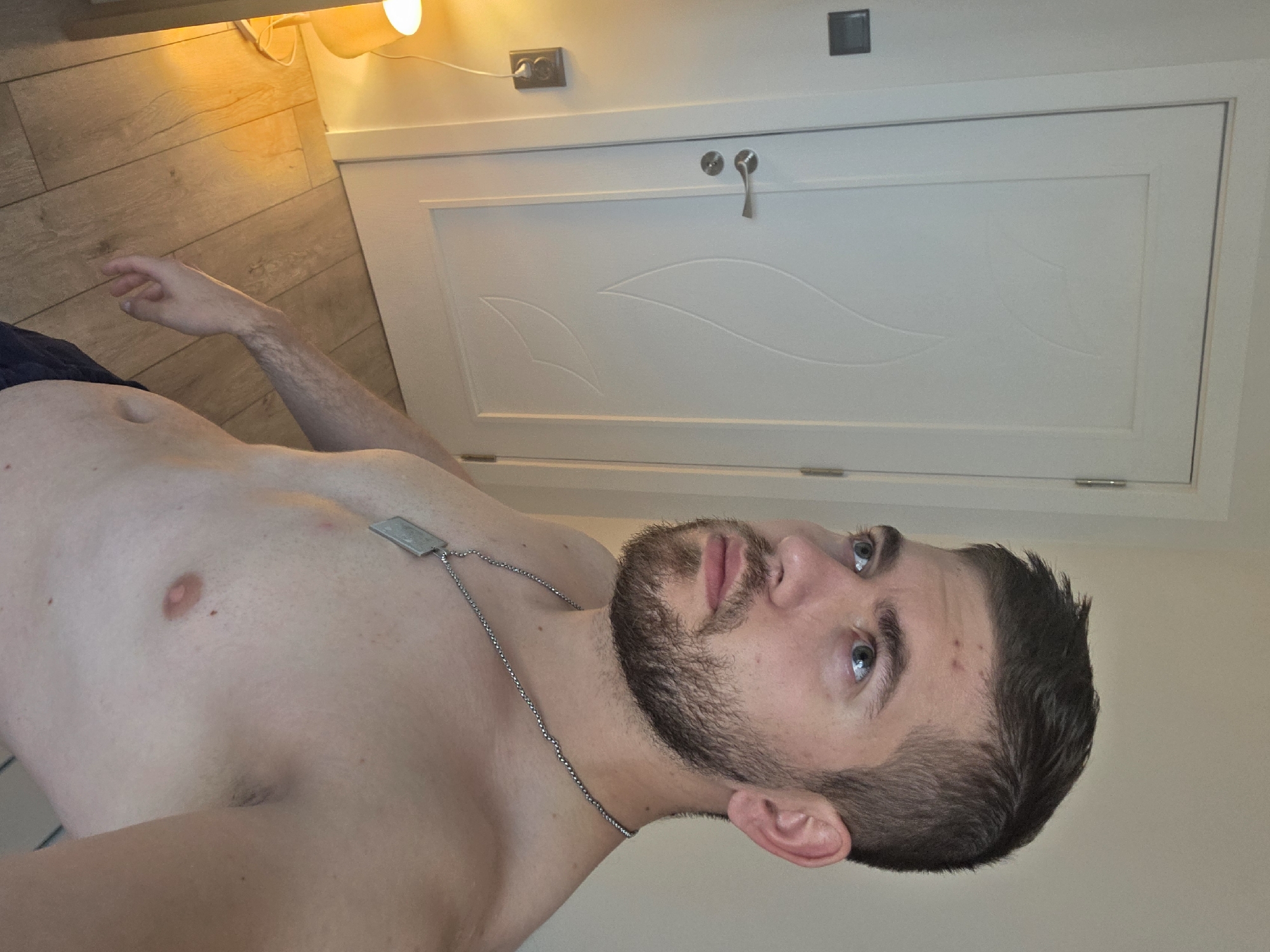 A medium shot depicts a fair-skinned man with a short, dark beard, gazing upward with light blue eyes. He is shirtless and wears a silver chain necklace with a rectangular pendant. The background features a white paneled door with silver hardware and a white wall to the right. The floor to the upper left is light wood. No landmarks associated with Yagodovo, Bulgaria, are visible.