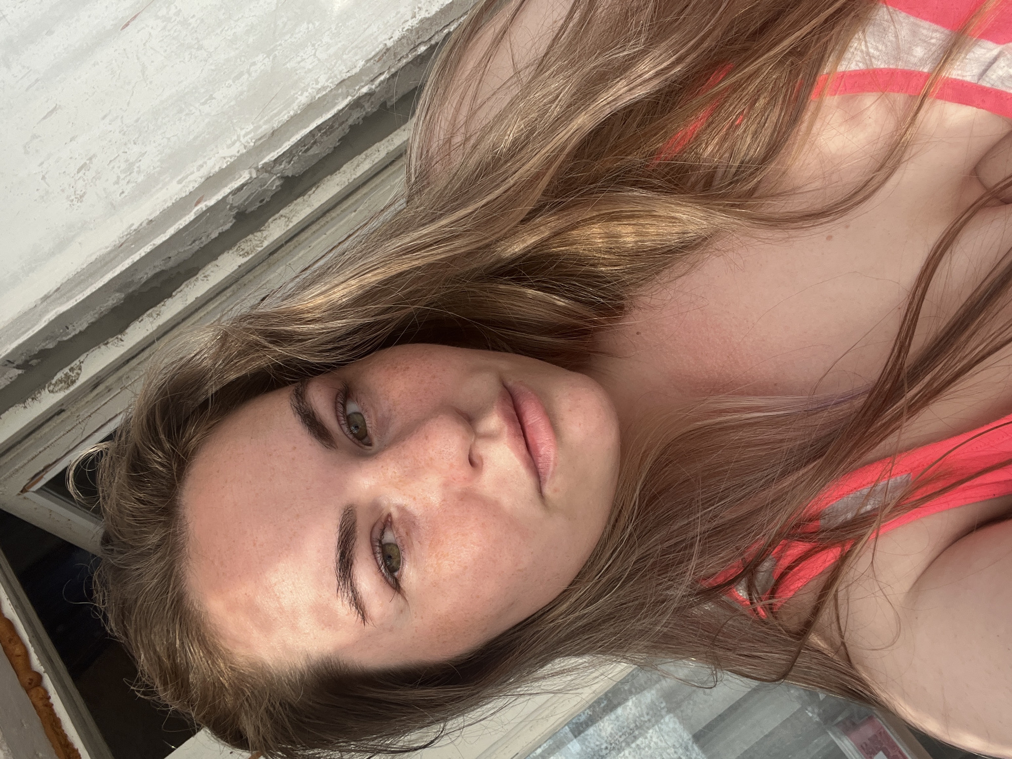 The image is a close-up, low-angle, selfie-style shot of a young Caucasian woman with long, wavy blonde hair. She is wearing a pink and white striped top. Her face is turned towards the camera, and she has fair skin with freckles. Her eyes are a light green, and her lips are slightly parted. Her eyebrows are dark and well-defined. The lighting suggests it is daytime, with sunlight casting shadows. The background appears to be a window frame, with a textured, off-white painted surface and some darkening on the lower edge. There are hints of a darker area visible through the window, possibly indicating an outdoor scene. The overall mood is relaxed and natural. No visible text is present in the image. The orientation of the image is rotated 90 degrees counter-clockwise.