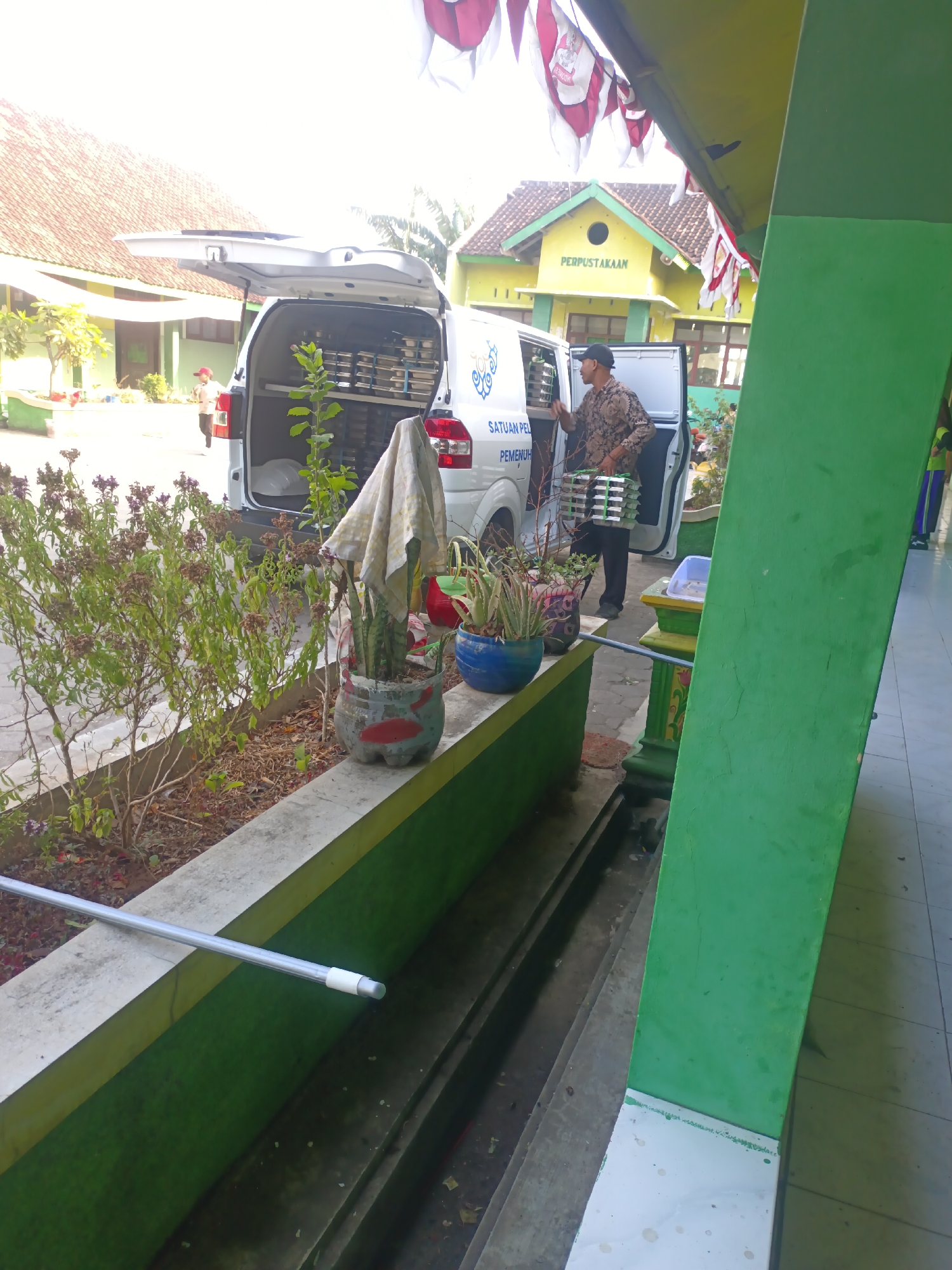 In Sarimulyo, Indonesia, a man is unloading stacked trays from the open back of a white van. The van has text on its side that reads "SATUAN PEL PEMENU". The man is wearing a patterned shirt and a dark cap, and he is holding a stack of trays. The van is parked in front of a building with a sign that says "PERPUSTAKAAN", indicating it is a library. There are red and white flags hanging from an overhang. In the foreground, there is a planter with plants, and a grey pole is visible. The ground is paved with tiles.