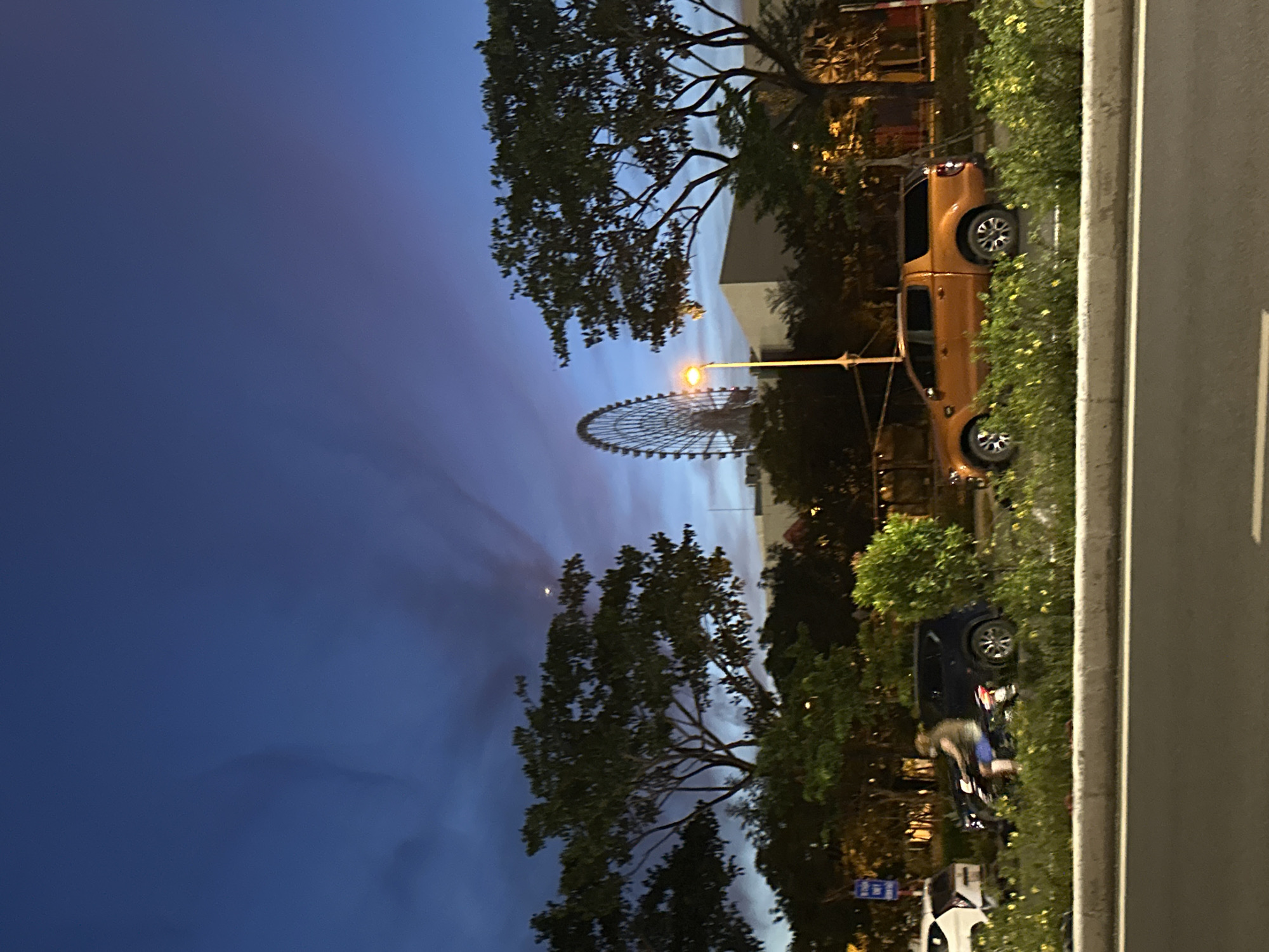 At twilight in Đà Nẵng, Vietnam, a street scene unfolds with a prominent Ferris wheel in the distance. The sky displays a gradient of deep blue to lighter hues with wispy clouds, and a bright celestial object, possibly Venus, is visible. Trees line the roadside, casting shadows and framing the view of urban structures. In the foreground, a dark-colored SUV is parked, and an orange pickup truck with a covered bed is also visible. A person is seen riding a motorcycle. A street lamp emits a warm glow. The overall setting is one of a transitioning day into night, with elements of leisure and transportation present.