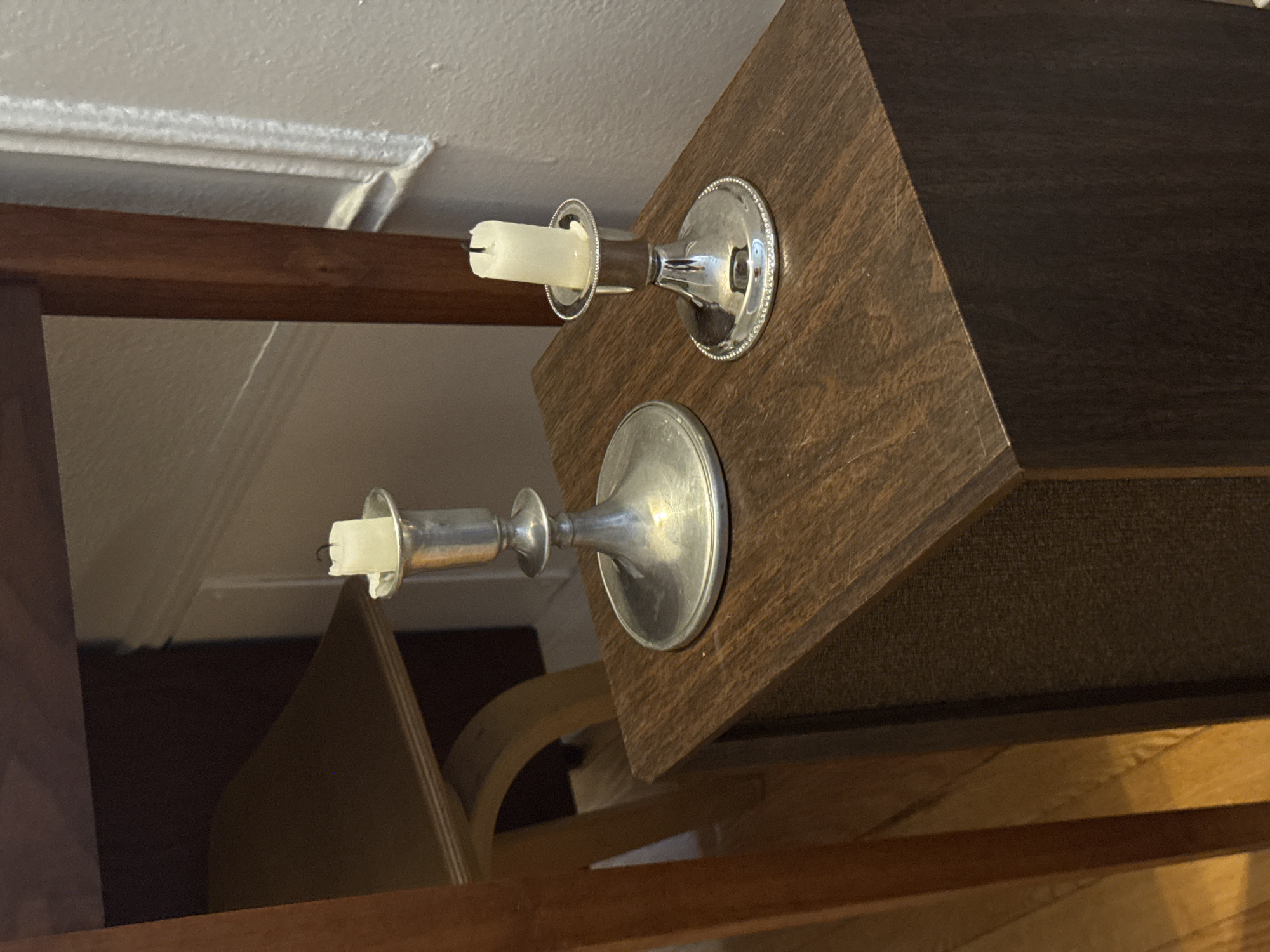 The image depicts two silver candle holders affixed to a dark-stained wooden structure. Each candle holder holds a white candle. In the background, architectural details of a room are visible, including molding and wooden furniture. The exact location in Montreal, Canada cannot be determined from this image.
