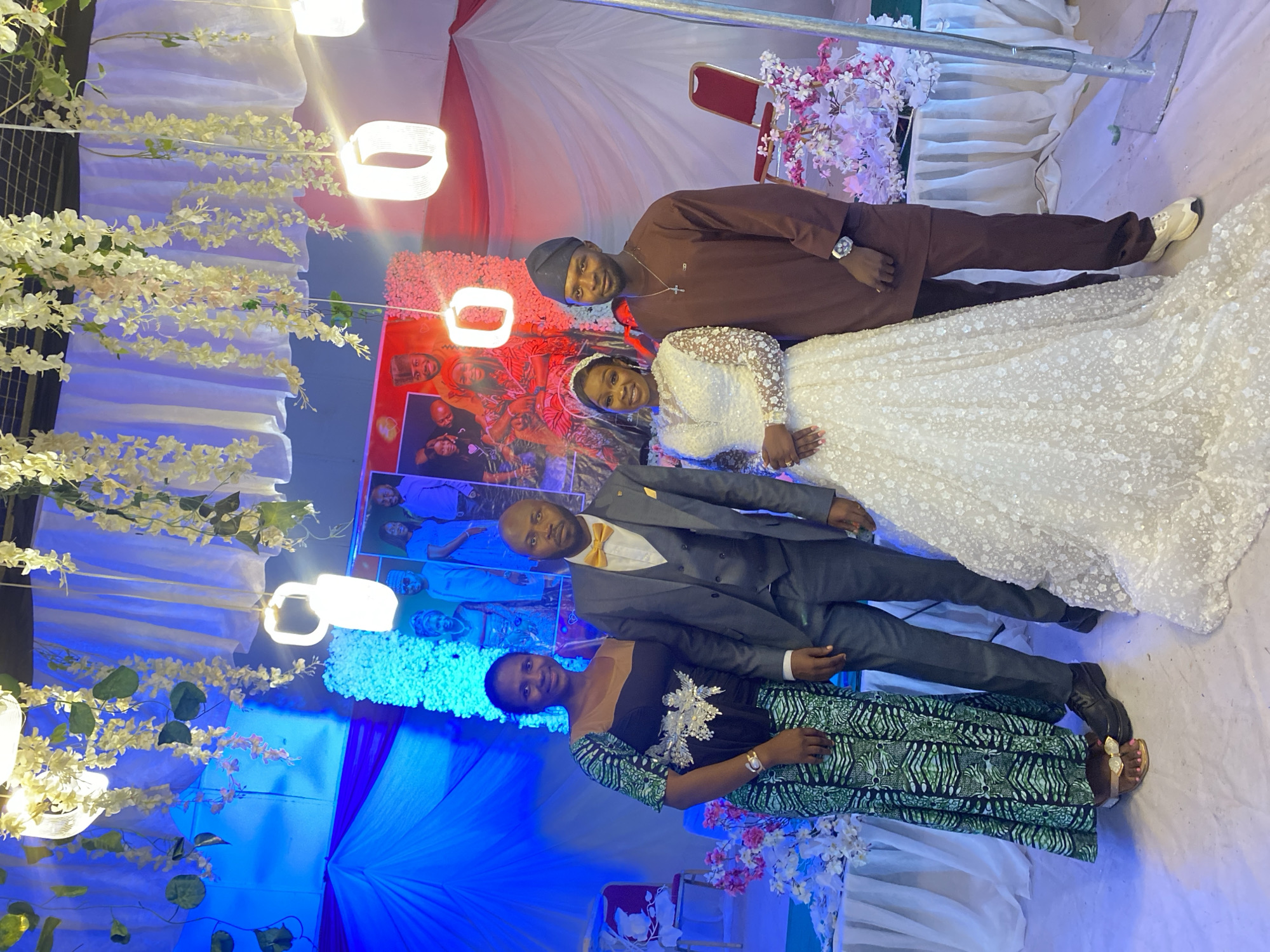 Four people are posing for a picture in what appears to be a wedding reception setting in Kuru Dungus, Nigeria. The bride is wearing a white, long-sleeved, beaded gown. A man in a brown tunic and cap stands beside her. Below the bride, a man in a grey suit and yellow bow tie stands with a woman in a dark dress with a green patterned overlay. The backdrop includes white and red draped fabric, flowers, and a framed photograph of the bride and groom. White floral decorations and square-shaped hanging lights are also visible.