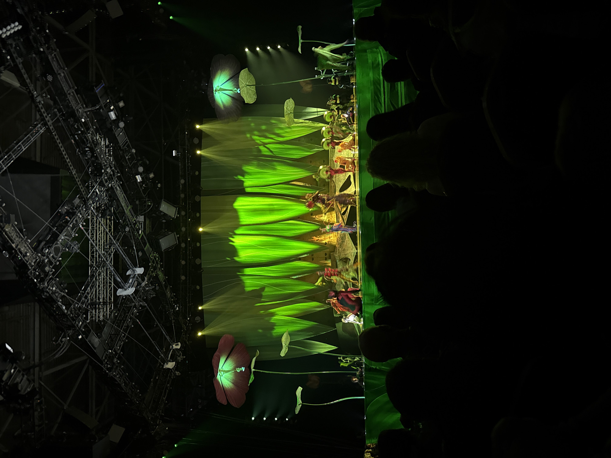 The image depicts a scene from a theatrical performance, possibly a play or musical, taking place indoors. On the left side of the image, there's a complex metal scaffolding structure with multiple lighting fixtures attached. The stage features a backdrop with green leaf-like projections illuminated by stage lights. There are performers on the stage, but they are somewhat blurred. The left side of the image is dark, suggesting the presence of an audience. There is no indication of the location being Sofia, Bulgaria, in the image.