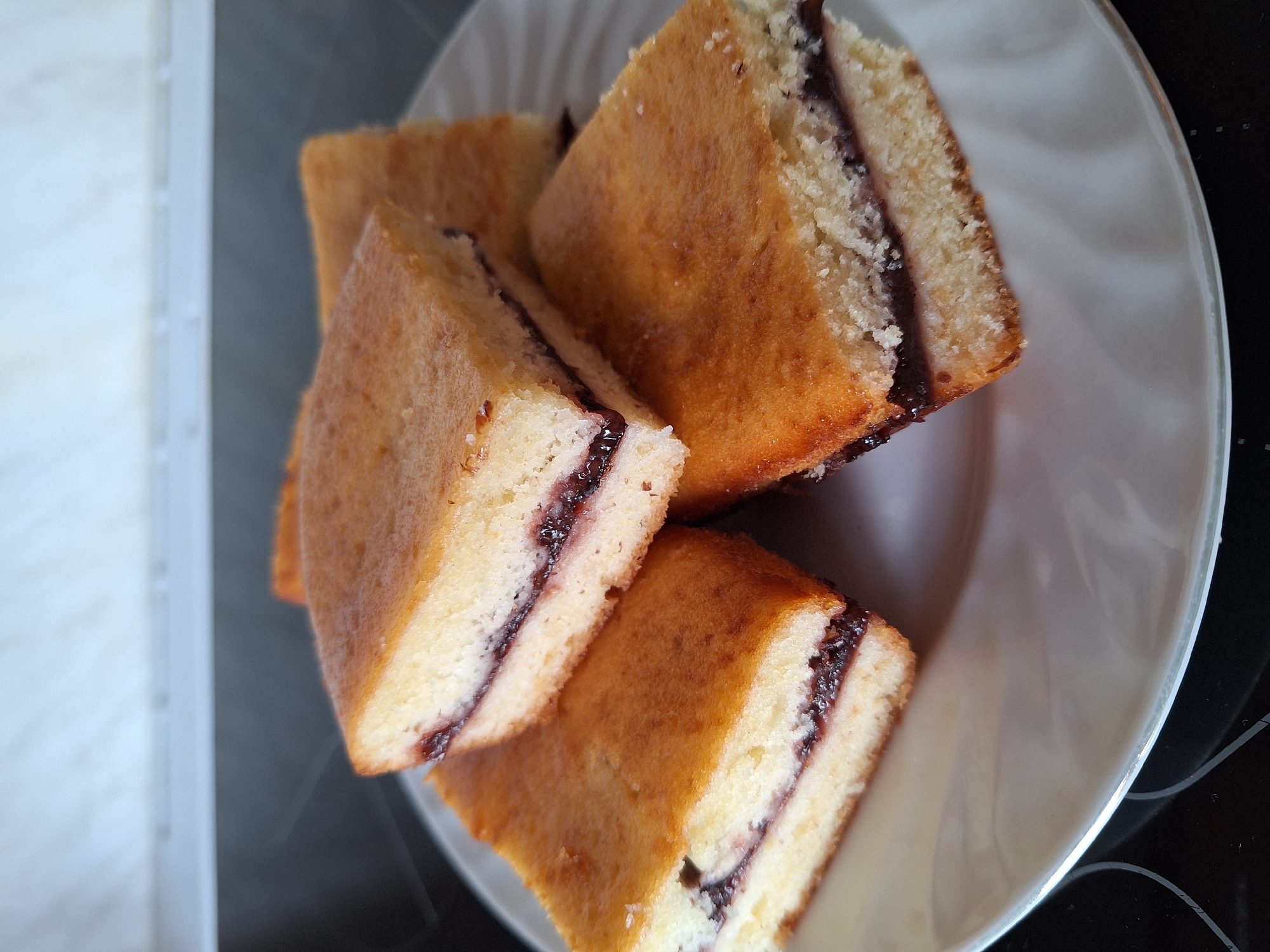 A high-angle, close-up shot shows four pieces of filled sponge cake on a white plate. The cakes are rectangular with a golden-brown crust and a light yellow interior, separated by a thin layer of dark filling. The cakes are stacked haphazardly on the plate. The plate appears to be on a dark surface, possibly a countertop. The location is unknown and cannot be determined from the image.