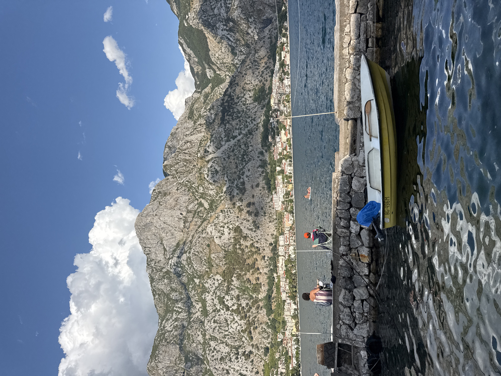In Prčanj, Montenegro, the image depicts a scenic bay with a stone embankment in the foreground. Two individuals are seated on chairs facing the water, with one person wearing a striped garment and the other a red cap. A yellow boat with a blue cover is moored to the embankment, displaying the registration "BG-496M". In the background, a town with white and red-roofed buildings is nestled at the base of a steep, rocky mountain. The water is calm, reflecting the sky and clouds, and several other people can be seen in the distance, some swimming or floating on the water. The sky is a clear blue with scattered white clouds.