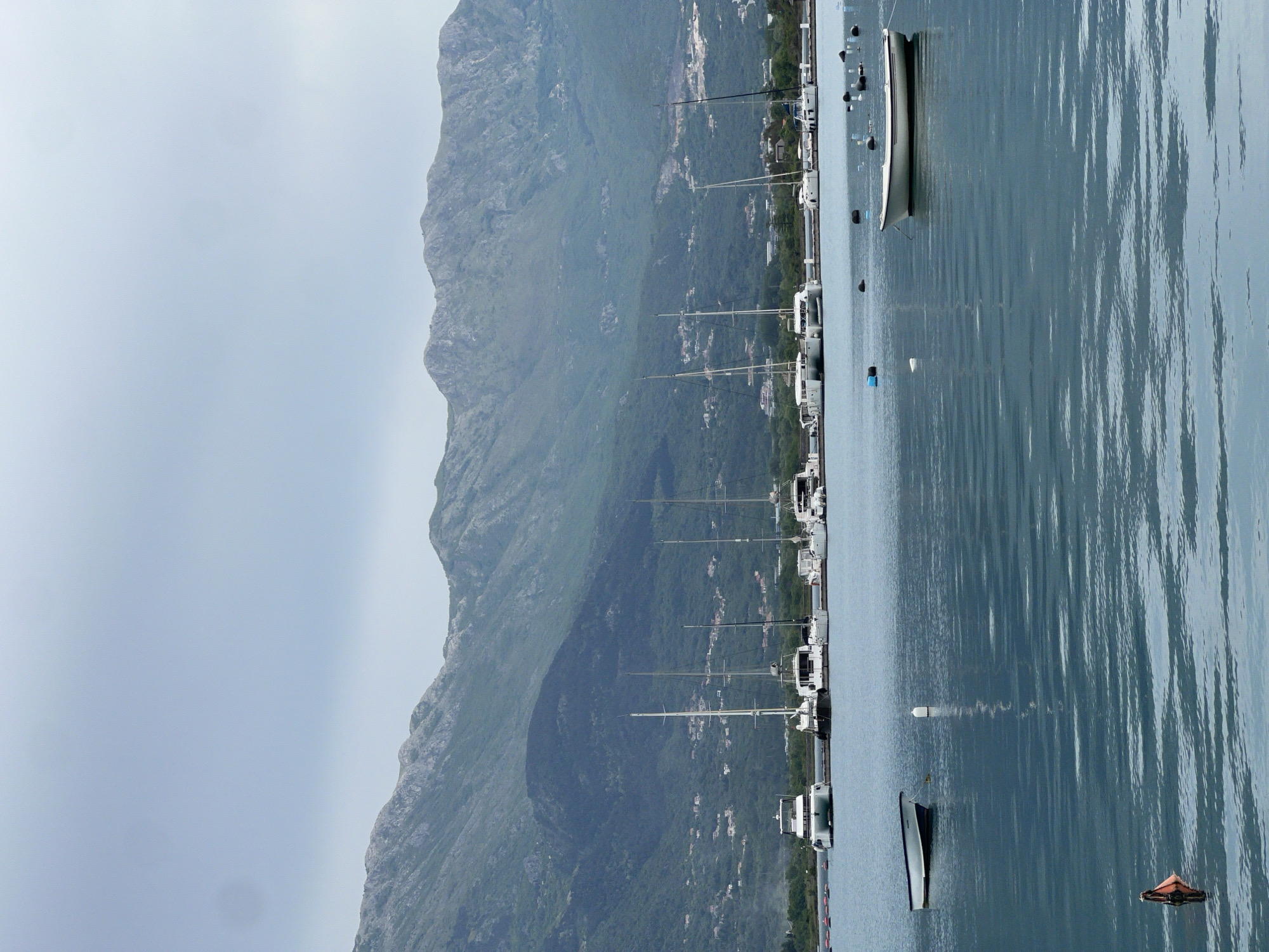 This image depicts a harbor in Đuraševići, Montenegro. Boats are moored along a dock on calm water. Mountains rise in the background, partially obscured by haze. The sky is overcast.