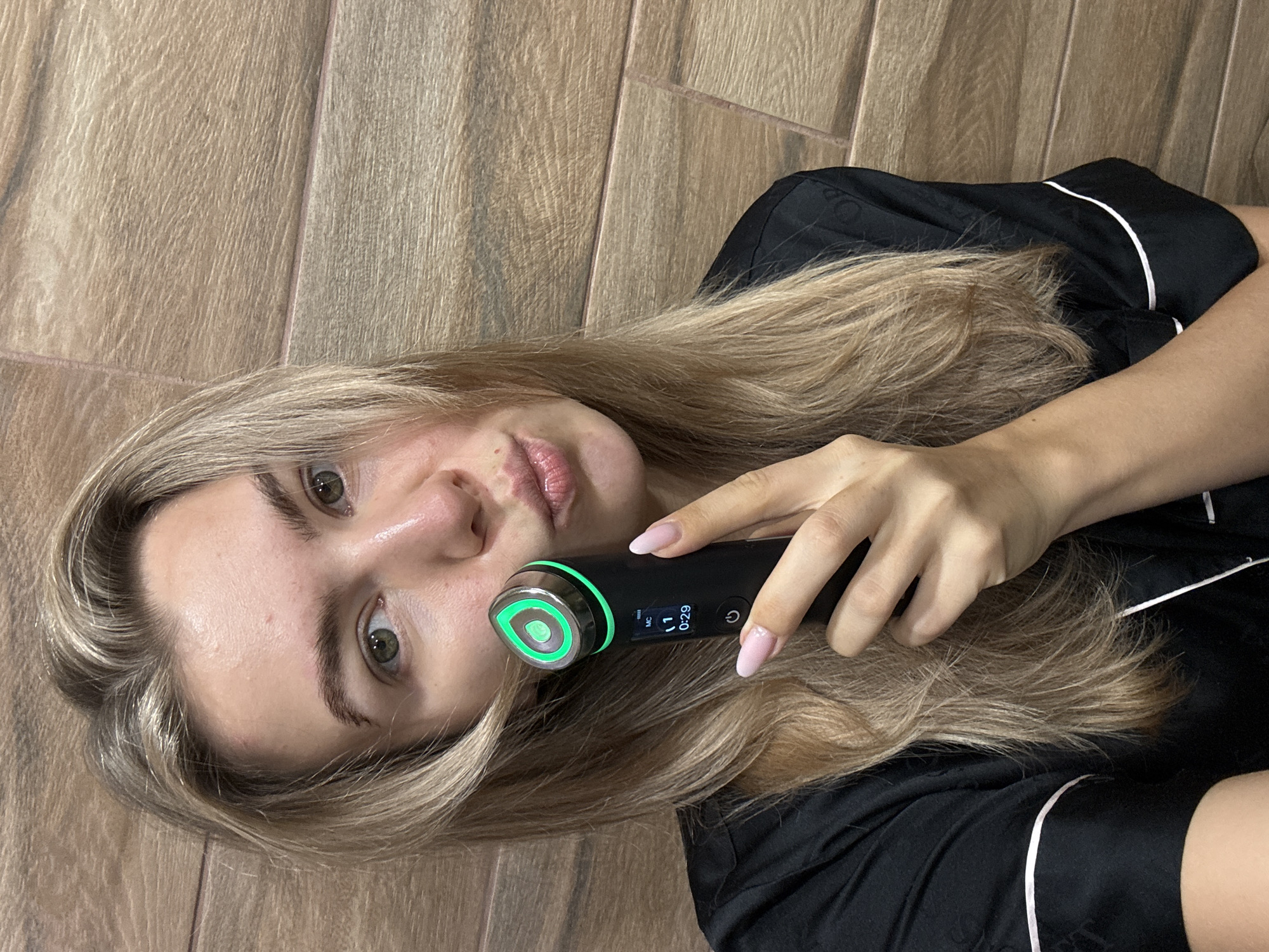 The image shows a young woman lying on her back, likely on a wooden floor. She is fair-skinned with blonde hair and is wearing a black pajama top with white piping. She is holding a black beauty device with a circular, glowing green light against her face. The device has a small screen displaying the numbers "0:29". Her fingernails are painted a light pink color. The background appears to be a wooden floor.