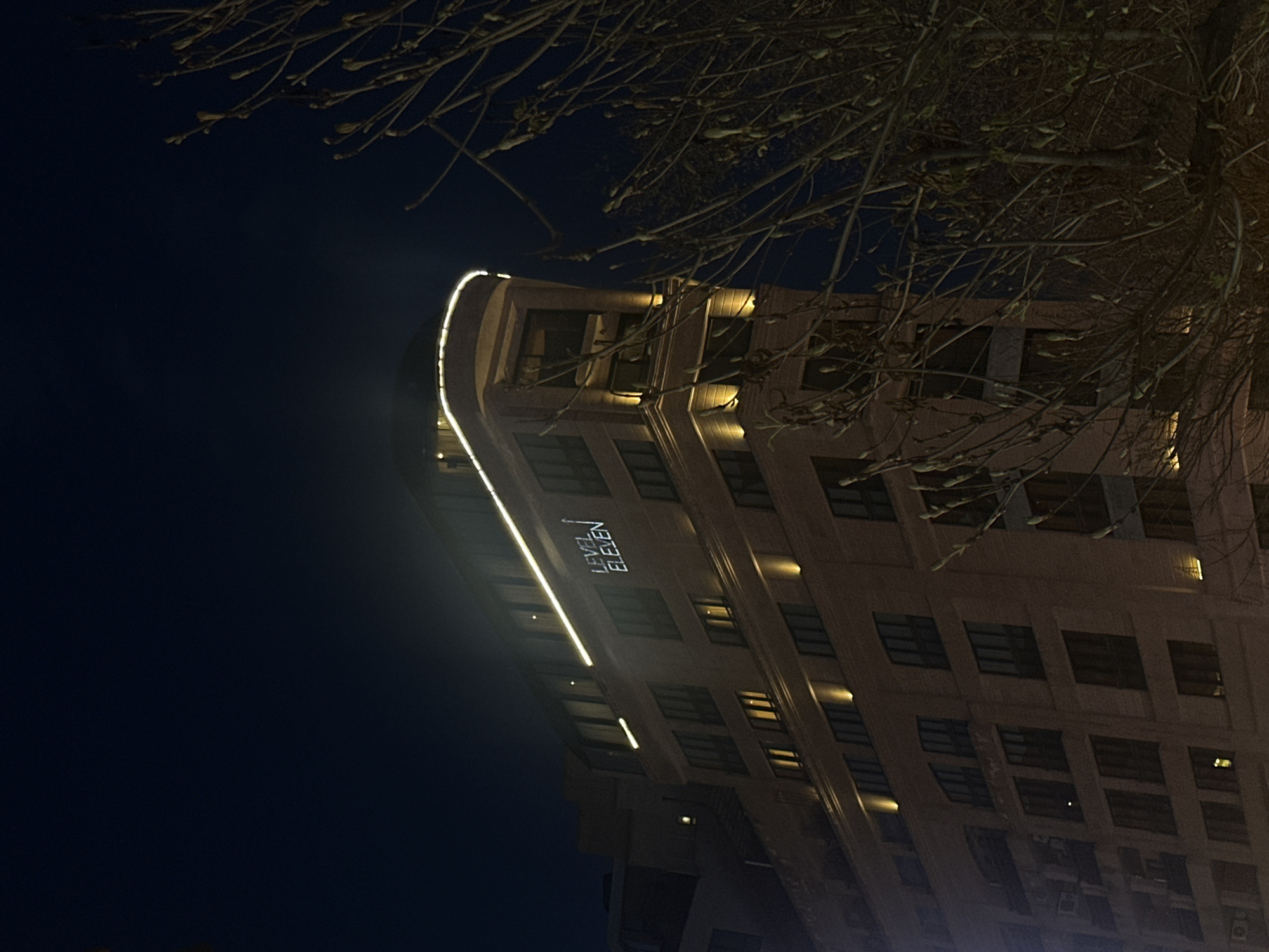 The image shows a building at night, likely in Yerevan, Armenia, given the text "LEVEL ELEVEN" on the building's façade, which corresponds to a hotel in that city. The building has a distinctive curved corner and is lit with a bright strip of light running vertically along the edge. The windows are rectangular and uniformly spaced. Tree branches are visible in the upper right corner and top center of the frame, partially obscuring the building and the dark sky. The overall light conditions are dim, with the building's illumination contrasting against the dark background.
