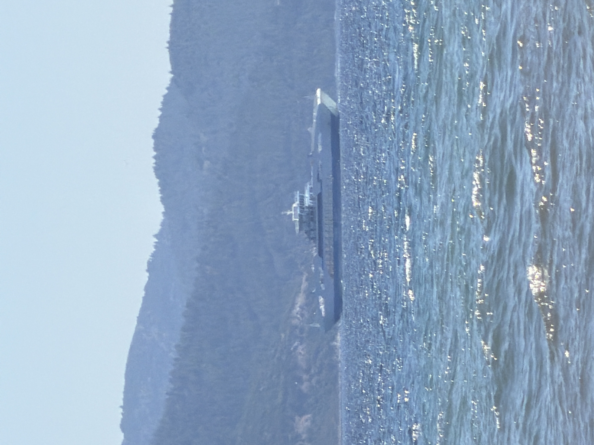 The image depicts a ferryboat navigating the waters near the coast of Keramoti, Greece. The ferry is the central object, seen from the side, with its layered decks and superstructure clearly visible. It appears to be in motion, indicated by the waves surrounding it and the slight tilt of the vessel. The water is a deep blue, reflecting the sunlight with shimmering highlights, suggesting it is daytime.
To the left of the ferry, a steep, tree-covered hillside rises, forming the coastline. The hillside is rendered in muted tones of green and blue, indicating distance and perhaps a hazy atmosphere. The sky above is a pale, clear blue, contributing to the overall serene and bright ambiance of the scene. The composition suggests a clear day with good visibility. There are no people clearly discernible on the ferry, and no text is visible in the image. The setting is clearly coastal, with the ferry acting as a mode of transport within a natural landscape.