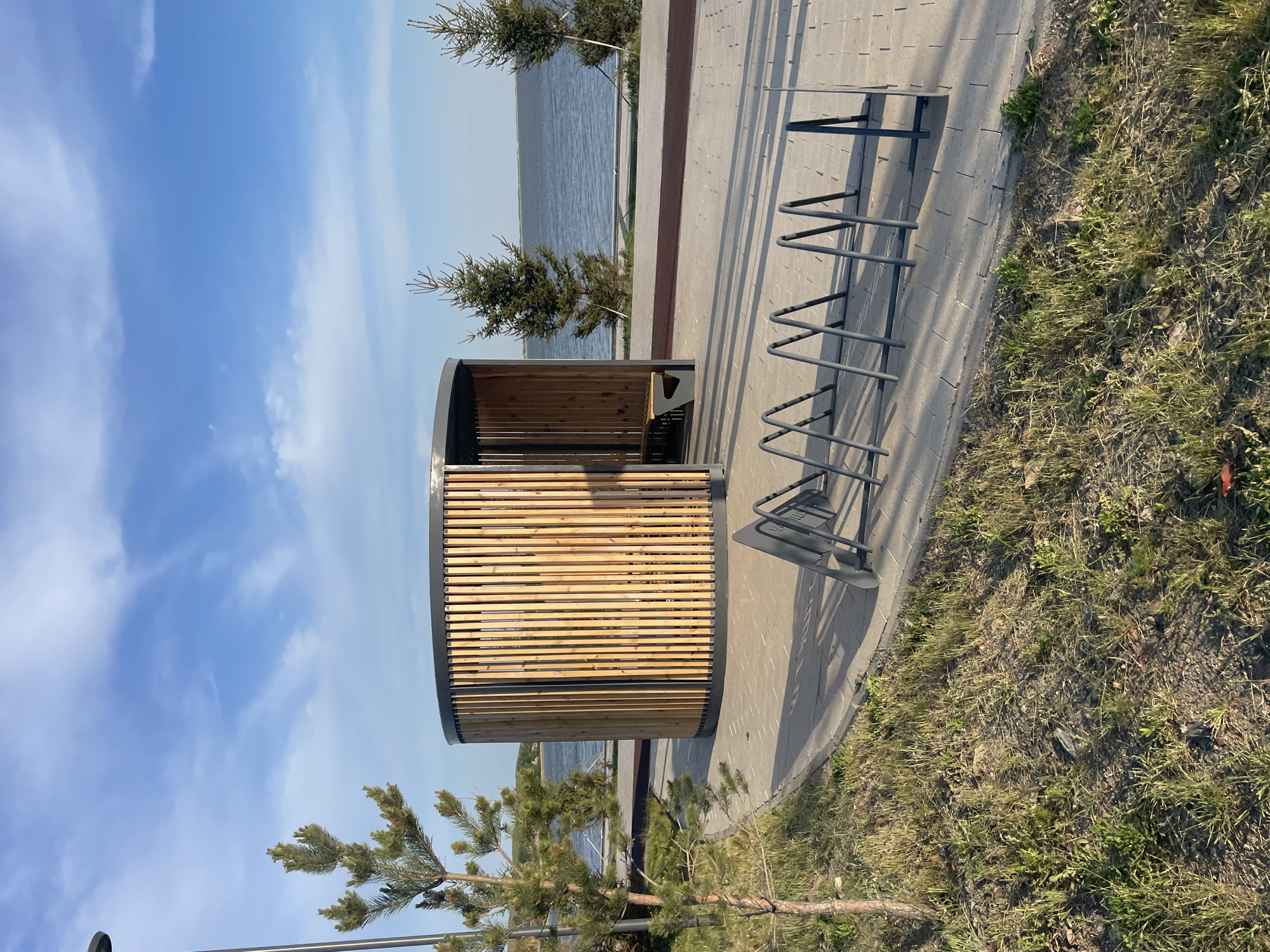 The image shows a rest area in what appears to be a park or recreational area, possibly in Kokshetau, Kazakhstan.
Key features:
* **Structure:** A semi-circular wooden structure with a curved roof. The structure provides seating and shelter.
* **Bicycle Rack:** A metal bicycle rack is situated on the right of the structure, indicating the location is intended to be bicycle-friendly.
* **Pavement:** The ground is paved with light gray concrete blocks.
* **Vegetation:** There is grass on the right. There are pine trees near the structure and in the background.
* **Sky and Water:** The sky is partially cloudy and a body of water is visible in the background, suggesting a lakeside or riverside location.
The scene is likely a public space intended for relaxation and recreation.