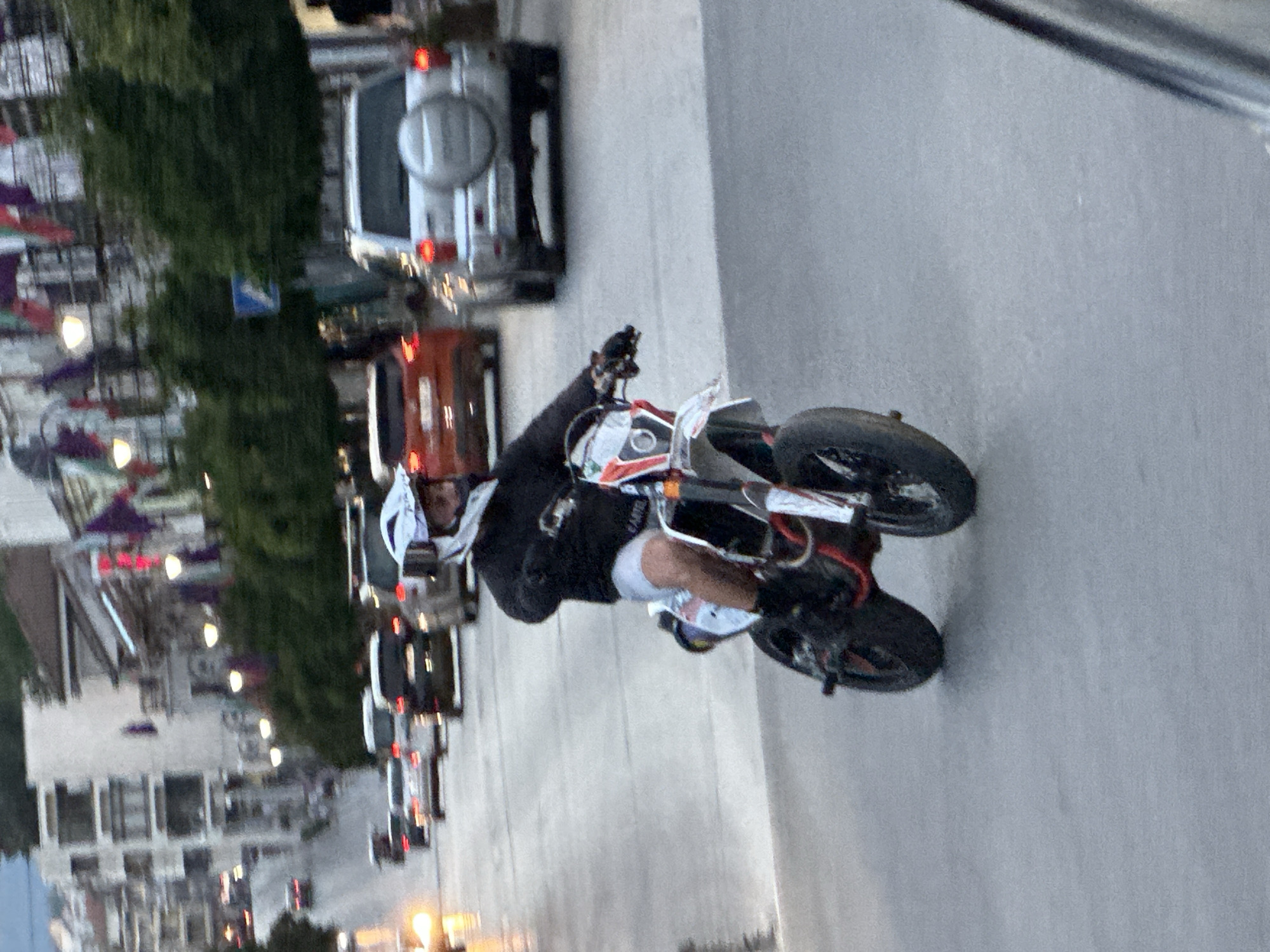 The image shows a person riding a dirt bike on a street in Veliko Tarnovo, Bulgaria. The rider is wearing a helmet, a black long-sleeved shirt, and shorts. The dirt bike is white with red and black accents. The street is paved and appears wet, suggesting it may have rained recently. In the background, there are several cars, buildings, and trees. The buildings are multi-story and have a light-colored facade. Flags or banners are visible hanging from some of the buildings. The overall scene suggests an urban environment with moderate traffic.