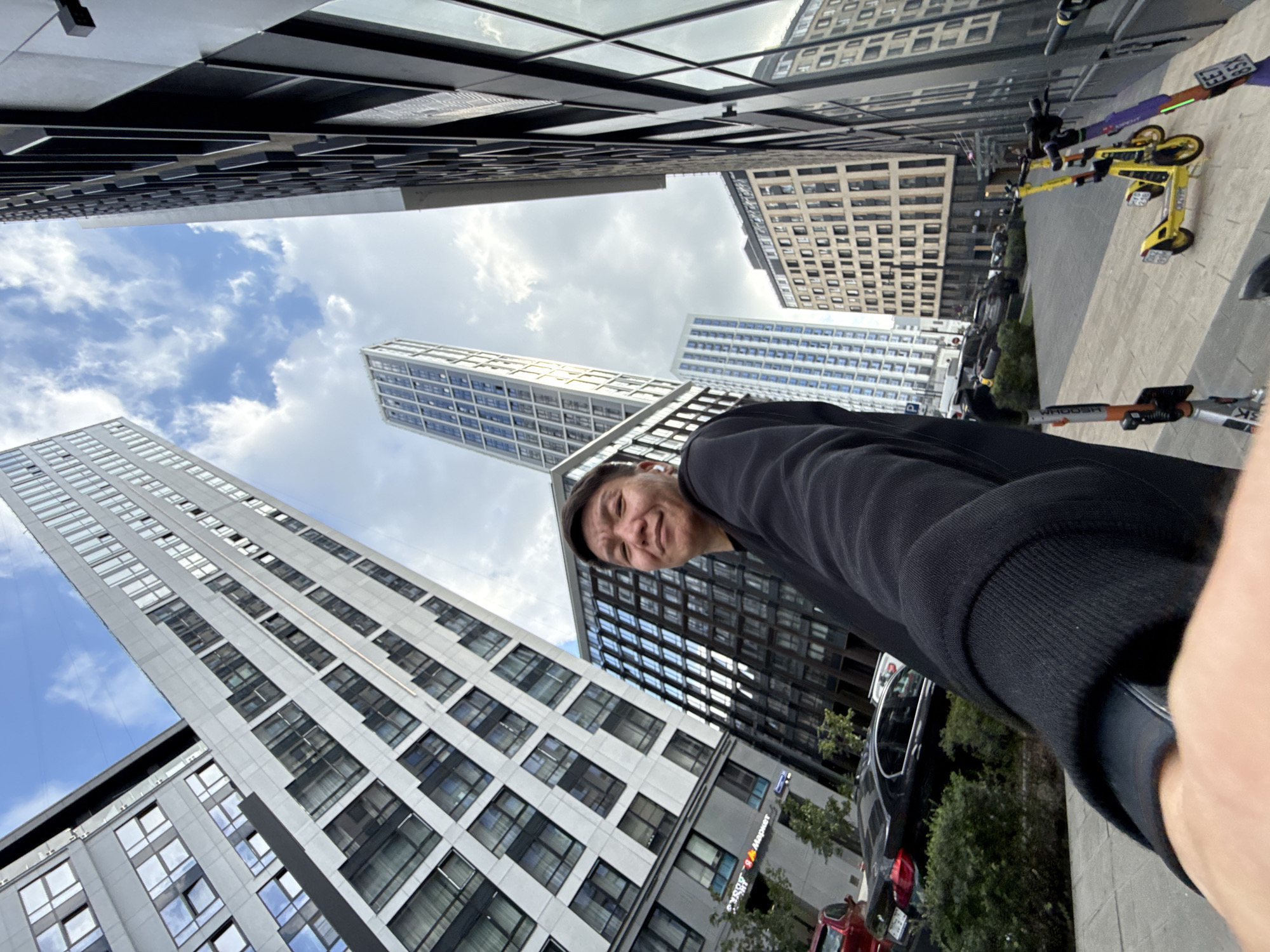 This is a first-person perspective, street-level, outdoor photograph taken in Moscow, Russia. The image captures a man's face and upper body as he is taking a selfie, with his left arm and hand extended towards the camera. He is wearing a black hooded sweatshirt and appears to have a smartwatch on his left wrist. The man is looking directly at the camera with a slight smile.
The background is dominated by modern, tall buildings with reflective windows and geometric architectural designs. One building on the left is white with many windows arranged in a grid pattern. Another building on the right is dark and sleek, also with numerous windows. In the distance, a very tall, slender skyscraper with blue-tinted windows stands out. The sky above is partly cloudy with a bright blue sky visible.
In the foreground, on the right side of the image, a row of parked electric scooters is visible on a paved walkway. Two prominent scooters are yellow and purple, with license plates. To the left of the scooters, a black car and a red car are parked on a street. Green bushes and trees are present in the middle ground, between the walkway and the cars. The overall impression is of a modern urban environment in Moscow.