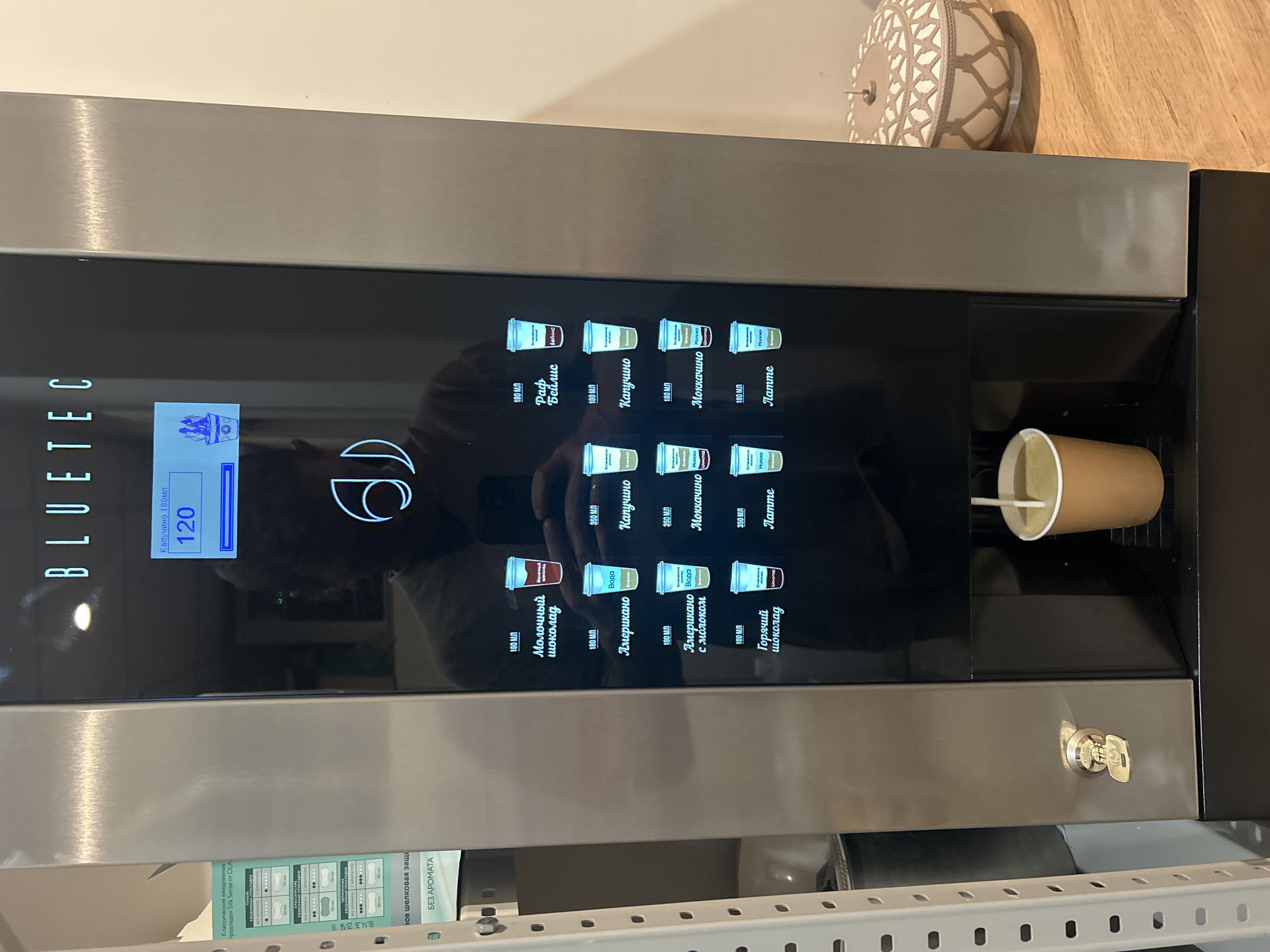 The image shows a BLUETEC beverage vending machine in what appears to be a public space. The machine has a digital display that shows "120" in large numbers, with the text "Капучино 180мл" above it. Below the display, there is a selection of drink options in Russian, each with an associated cup icon. Some of the drink names visible include "Молочный шоколад", "Американо", "Капучино", "Моккачино", and "Латте". A cup is currently being filled with a light-colored liquid from the dispenser. To the left of the machine, a white metal shelving unit is visible, with some printed material attached to it. The machine is situated on a wooden surface, and a decorative bowl is visible to the right. There is no specific information or visual cues in the image that indicate the location is Postol, Russia.