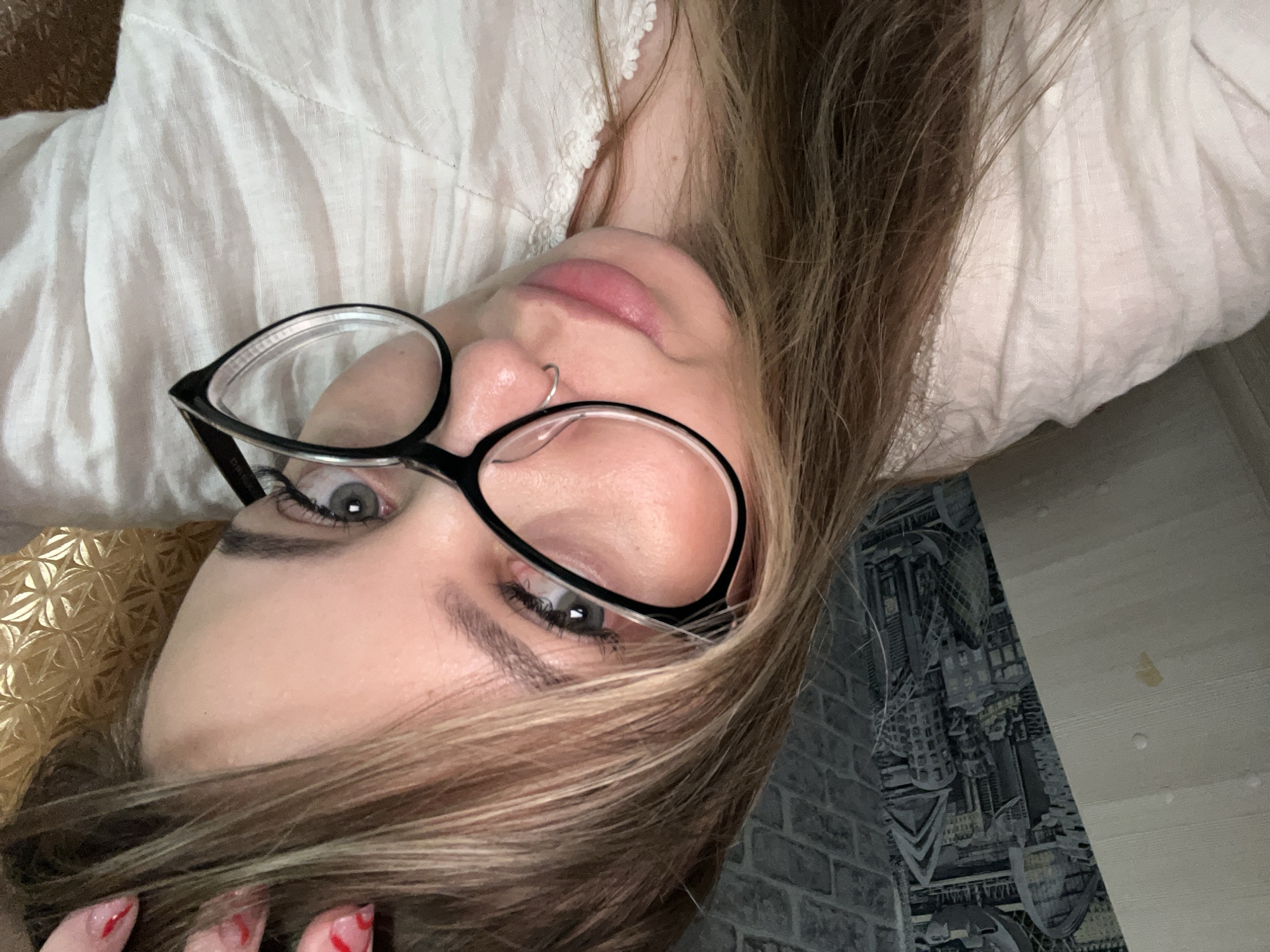 A young woman with light brown hair and grey eyes, wearing black-rimmed glasses and a nose ring, is lying down, looking up and towards the viewer. She is wearing a white, possibly linen, top. Part of her hand, with red nail polish, is visible in the lower left corner. The background includes a gold-colored patterned object behind her head and a patterned wall, possibly wallpaper, resembling brick and cityscapes. The location in the city of Novopokrovskaya, Russia is not deducible from the image.