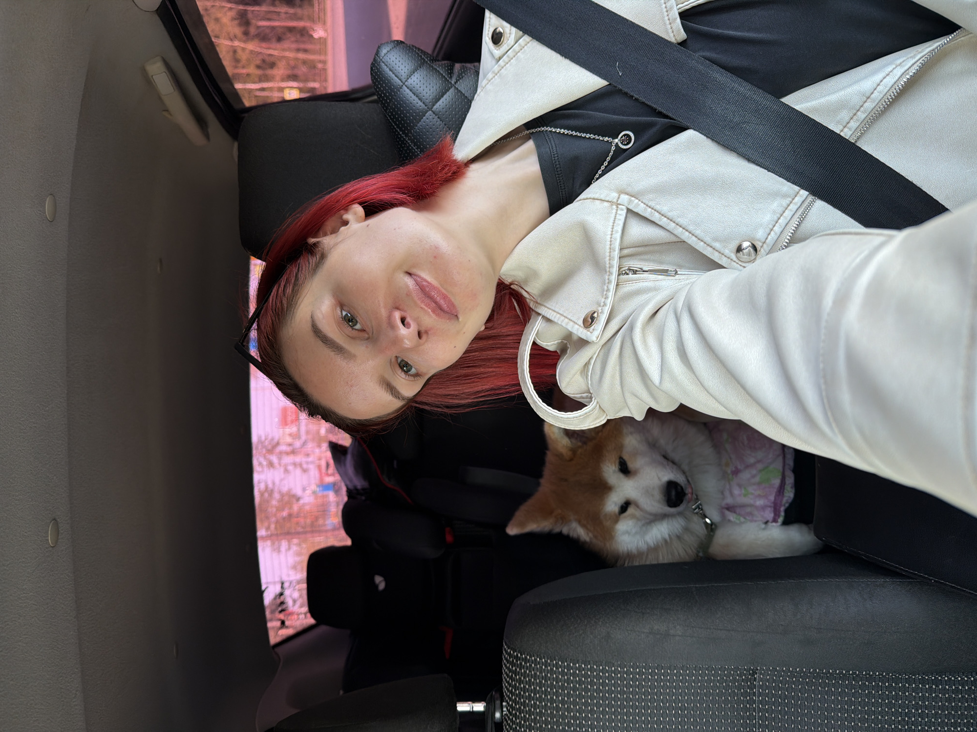 A woman with red hair is wearing a white jacket, a seatbelt, and black glasses. She is taking a selfie in what appears to be the front seat of a vehicle. Next to her is a dog, wearing a purple patterned vest, sitting in the passenger seat. The image was taken in Chaikovsky, Russia. The background shows an out-of-focus view of buildings, likely taken from the car's window.