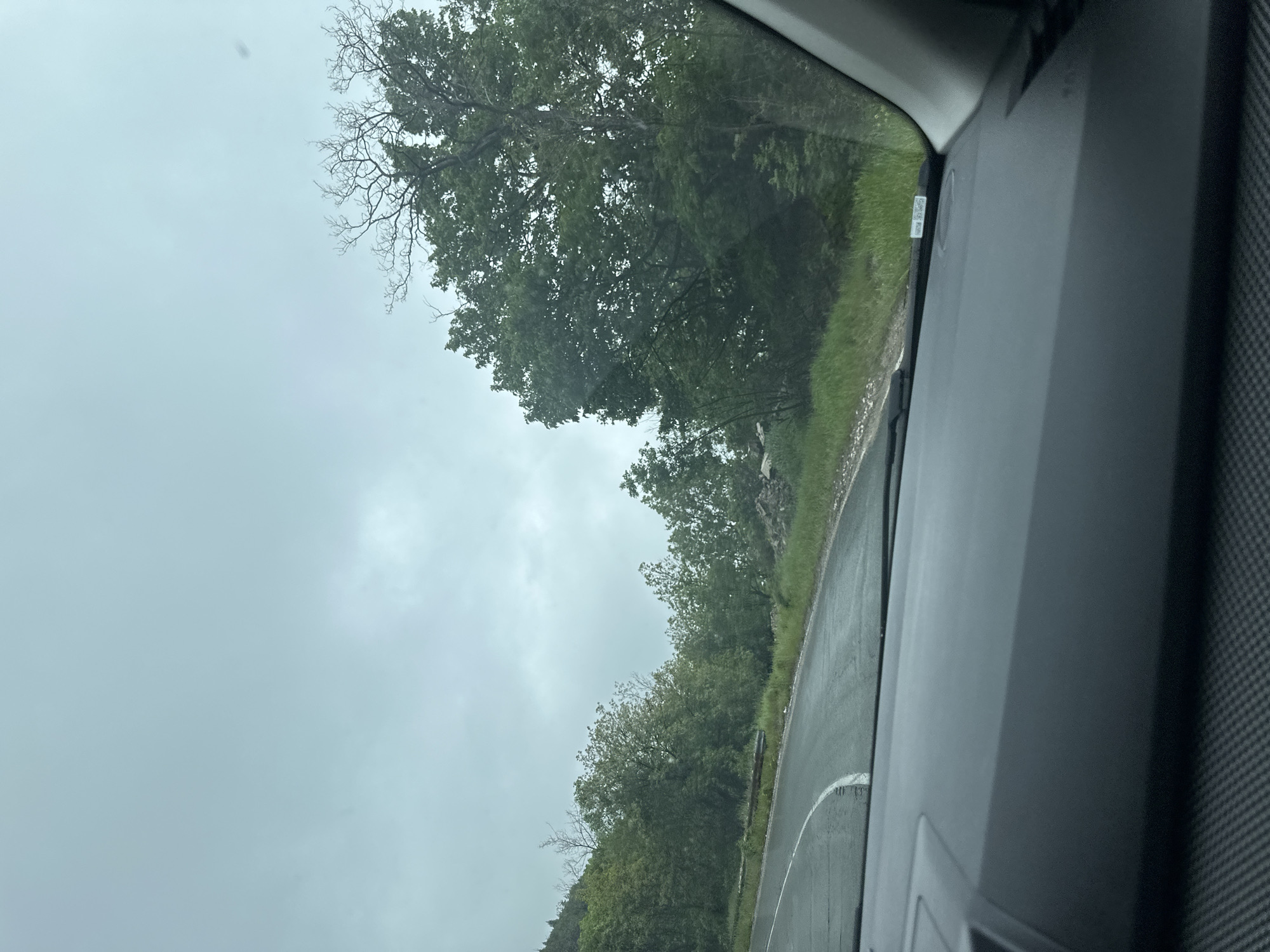 The image shows a view from inside a vehicle driving along a road in an area with trees and overcast skies. The road is wet, and a white lane marker is visible. The foreground includes a section of the vehicle's interior. The specific location in Karnare, Bulgaria cannot be determined from this image.