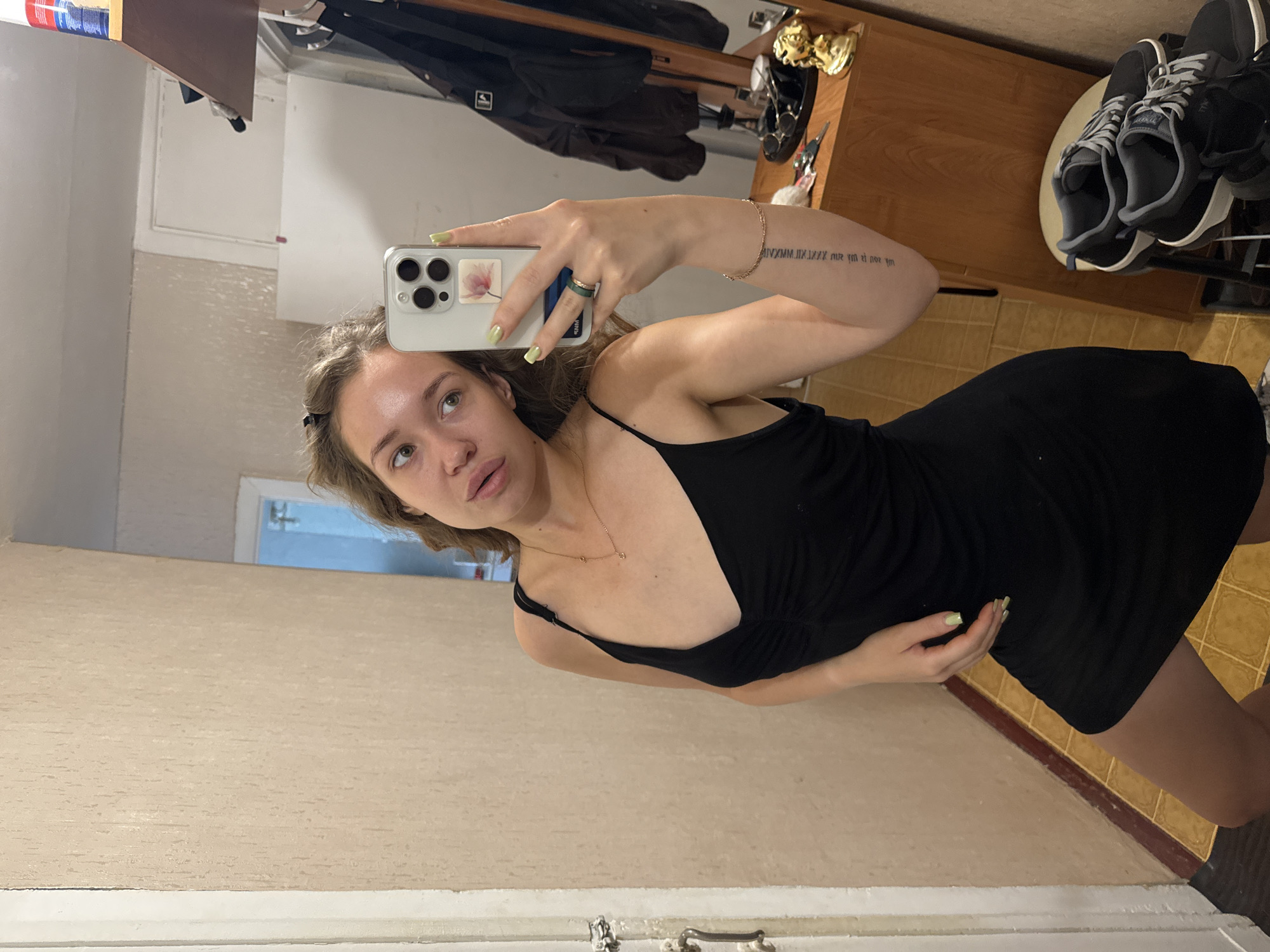 A young woman with light brown wavy hair is taking a mirror selfie in what appears to be a hallway or dressing area. She is wearing a black slip dress with thin straps and a V-neckline. Her right arm is raised, holding a smartphone. She has light green nail polish on her fingernails. A tattoo is visible on her left forearm. She is wearing a gold necklace with small pendants. On a wooden dresser in the background, there are some personal items, including sunglasses and a small golden figurine. A pair of sneakers are placed on a stool to the right of the dresser. The walls are textured and painted a neutral color. The floor is tiled in a yellowish-brown pattern. The overall impression is of a casual, personal space within a residential setting.