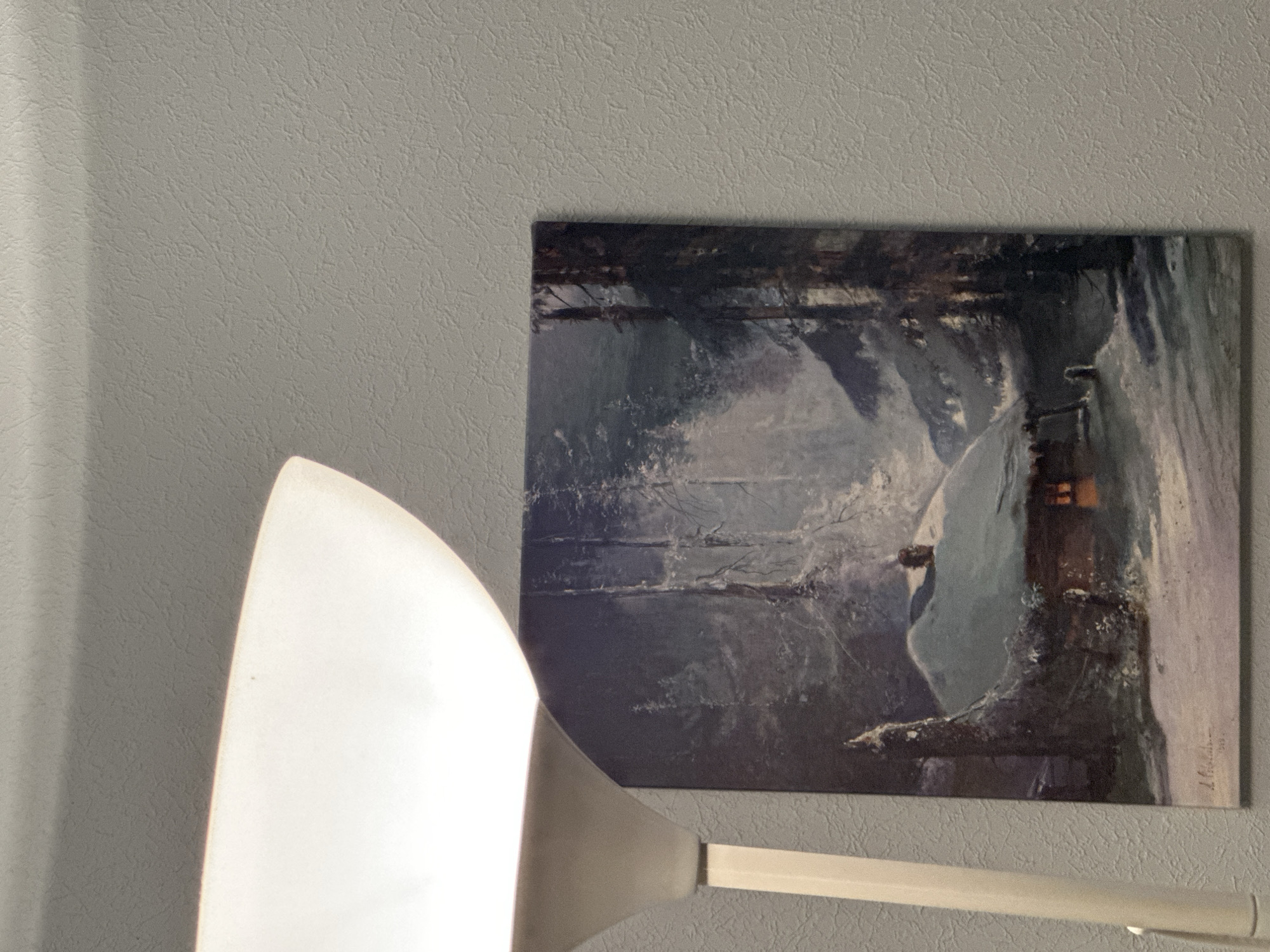 The image shows a painting of a snow-covered cabin in a forest, partially illuminated by a warm light from its windows. A white floor lamp is positioned to the left of the painting, casting a soft glow on the wall. The painting is signed in the lower left corner with what appears to be "J. Holsen" and dated "1919". The background of the painting depicts a dark, dense forest with snow-laden trees. The wall behind the painting has a textured finish, and a shadow from the lamp is visible on the upper portion of the wall. The scene appears to be indoors, given the presence of the lamp and the painting on the wall. The location in the city of Nevinnomyssk, Russia, is not discernible from the visual information provided in the image.