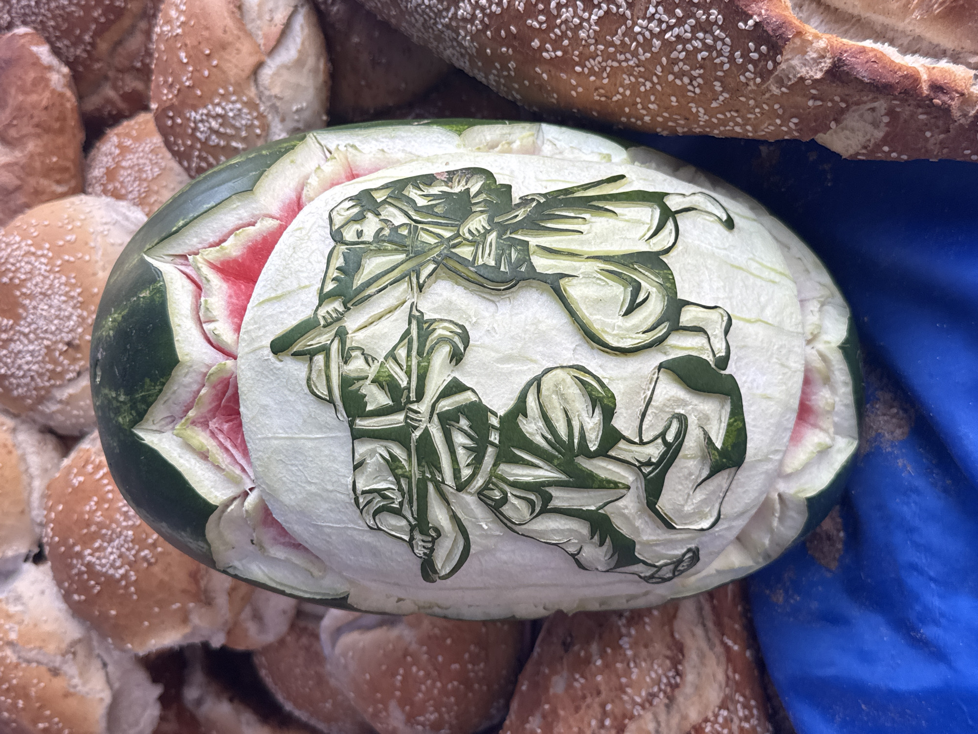 A high-angle, close-up shot shows a carved watermelon set amid a pile of bread and a blue cloth. The watermelon is oval-shaped, with the dark green outer rind partially removed to reveal the white flesh underneath. Carved into the white flesh is a scene depicting two figures with rifles. The remaining outer rind around the carved area is partially removed in a decorative, petal-like pattern, revealing the red flesh of the watermelon. The bread surrounding the watermelon appears to be small, round rolls, dusted with white sesame seeds. A brown loaf of bread, also covered in sesame seeds, rests near the top right corner of the image, and a blue cloth is partially visible on the right side. This type of watermelon carving is commonly sold in markets in Puebla, Mexico.