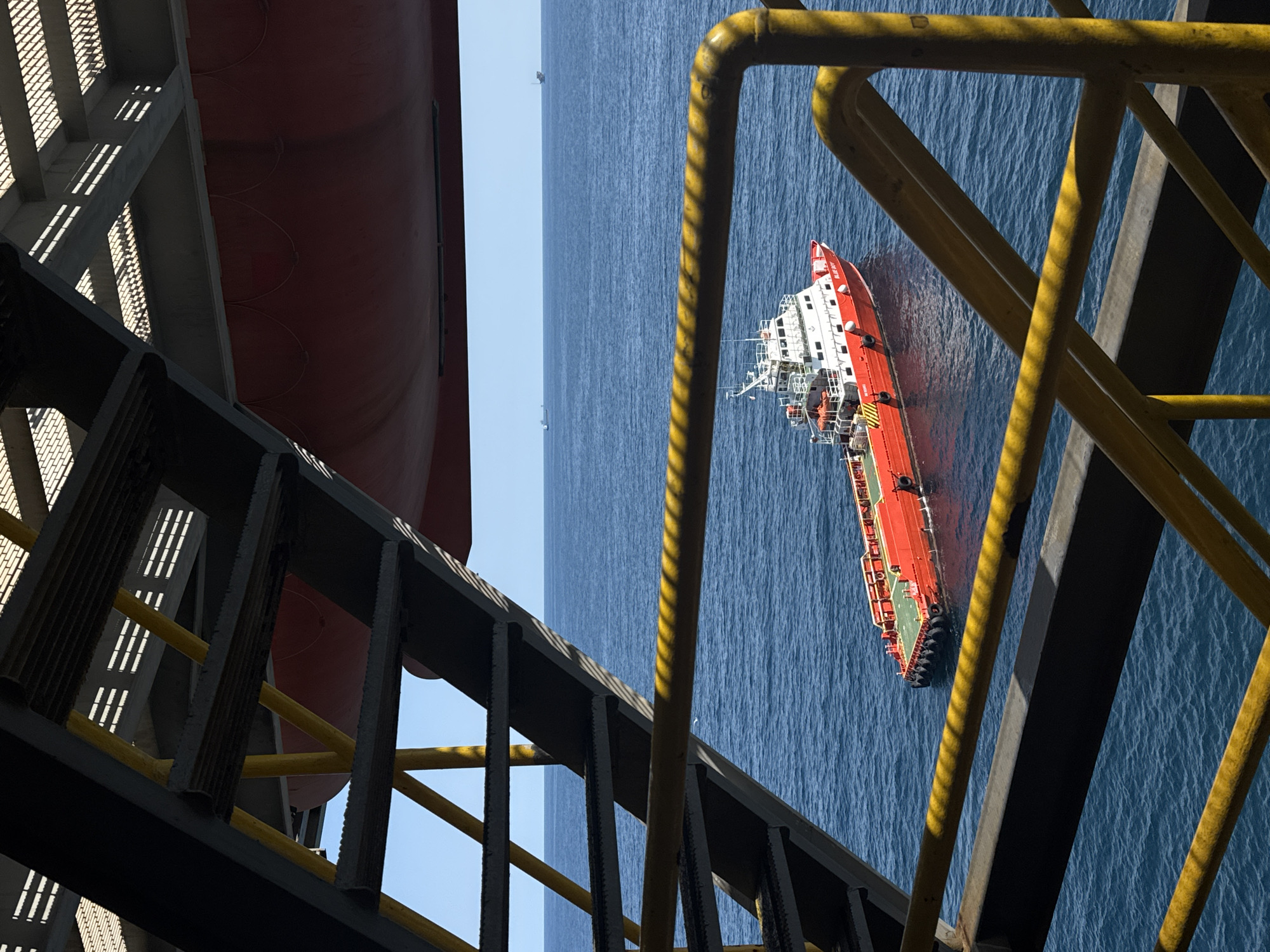 In an offshore setting, a red and white boat is visible in the blue ocean. The boat appears to be a supply vessel, with a flat deck and a superstructure. In the foreground, metal stairs and railings, painted yellow and black, lead down towards the water. The stairs and railings are part of a larger structure, possibly an oil rig or a platform. The sky is clear and blue, suggesting a sunny day. In the distance, other vessels and structures can be seen on the horizon. The overall scene depicts a working environment in the open sea.