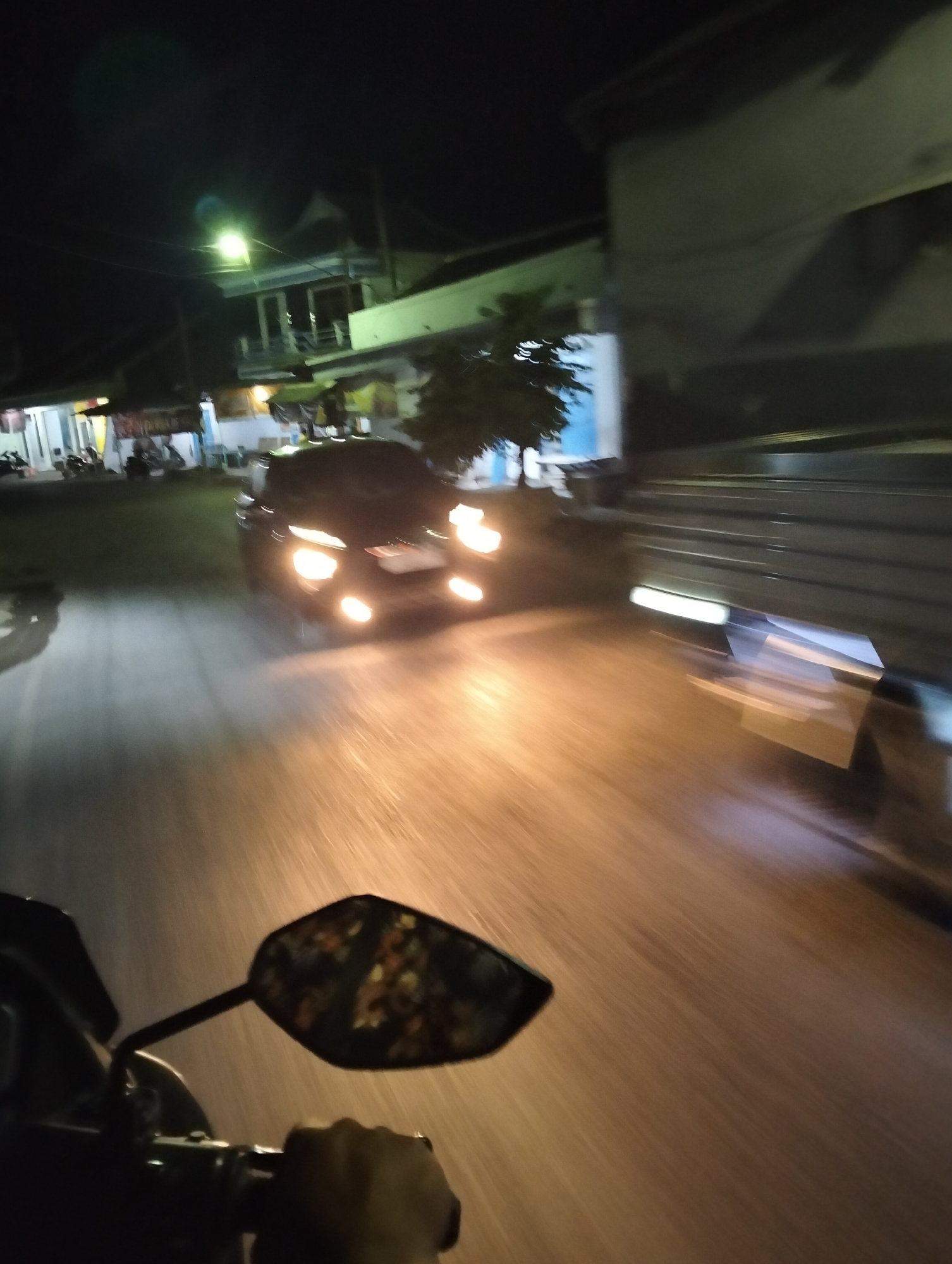 The image depicts a street scene at night in Muncar, Indonesia, likely captured from a moving vehicle. In the foreground, a hand is visible gripping a handlebar, and a motorcycle mirror reflects a colorful, abstract pattern, suggesting the presence of decorative items or reflections from nearby lights. The road ahead is illuminated by headlights of a dark-colored car, which is blurred due to motion. The car's headlights are prominently lit, casting a warm glow on the wet or reflective road surface. To the right, a large, dark structure, possibly a truck or a building with corrugated metal siding, dominates the scene. Buildings with lit windows and signs are visible on the left side of the street, indicating a commercial or residential area. A street light with a greenish hue illuminates a portion of the background. The overall impression is one of motion and a lively night atmosphere in a populated area of Muncar.