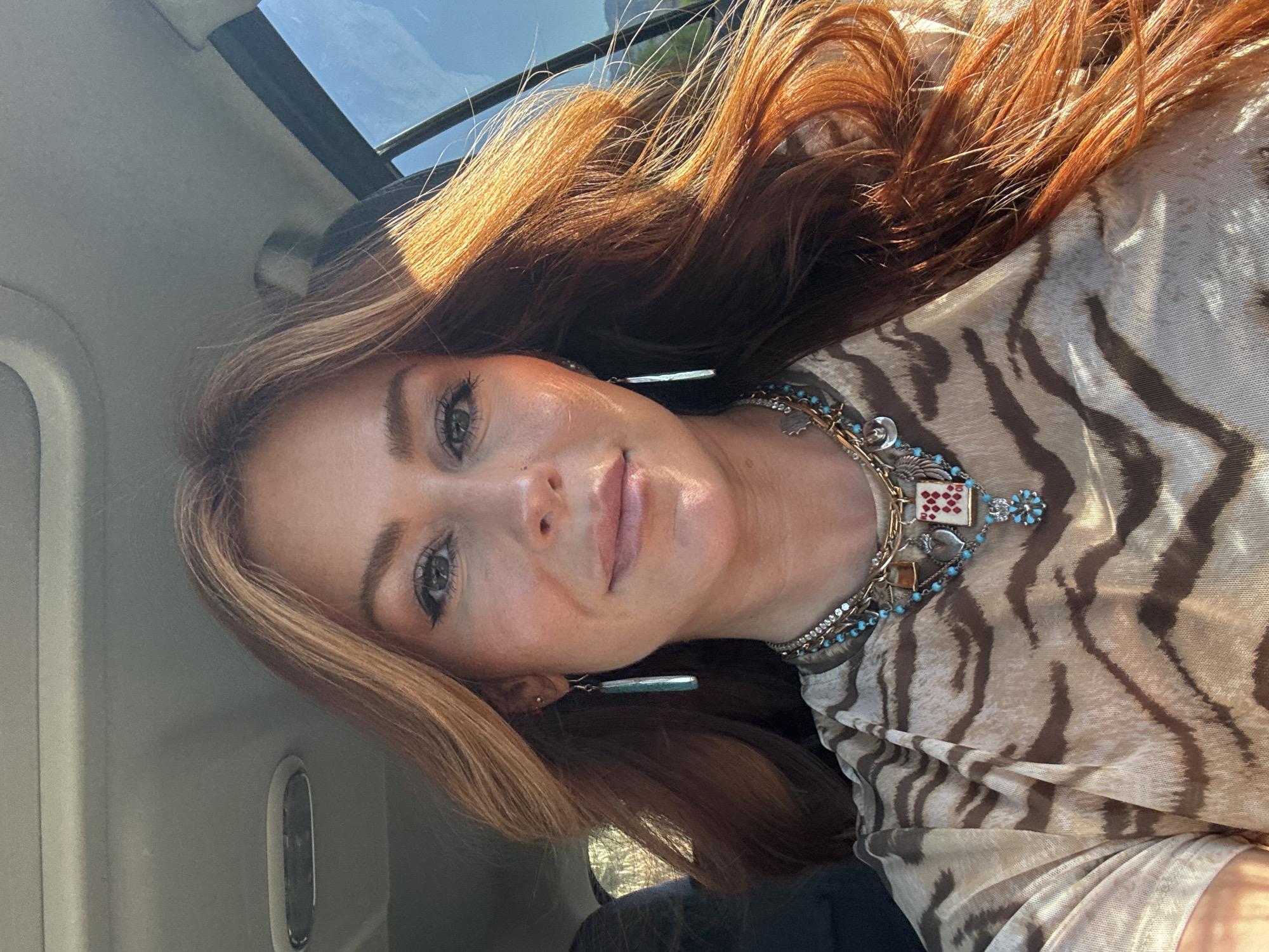 A woman with reddish-brown hair and light skin is taking a selfie inside a car, likely in La Quinta, United States of America, as suggested by the environment. The woman is wearing a tiger-striped shirt and a necklace with charms. The car's sunroof is visible in the top left of the frame.