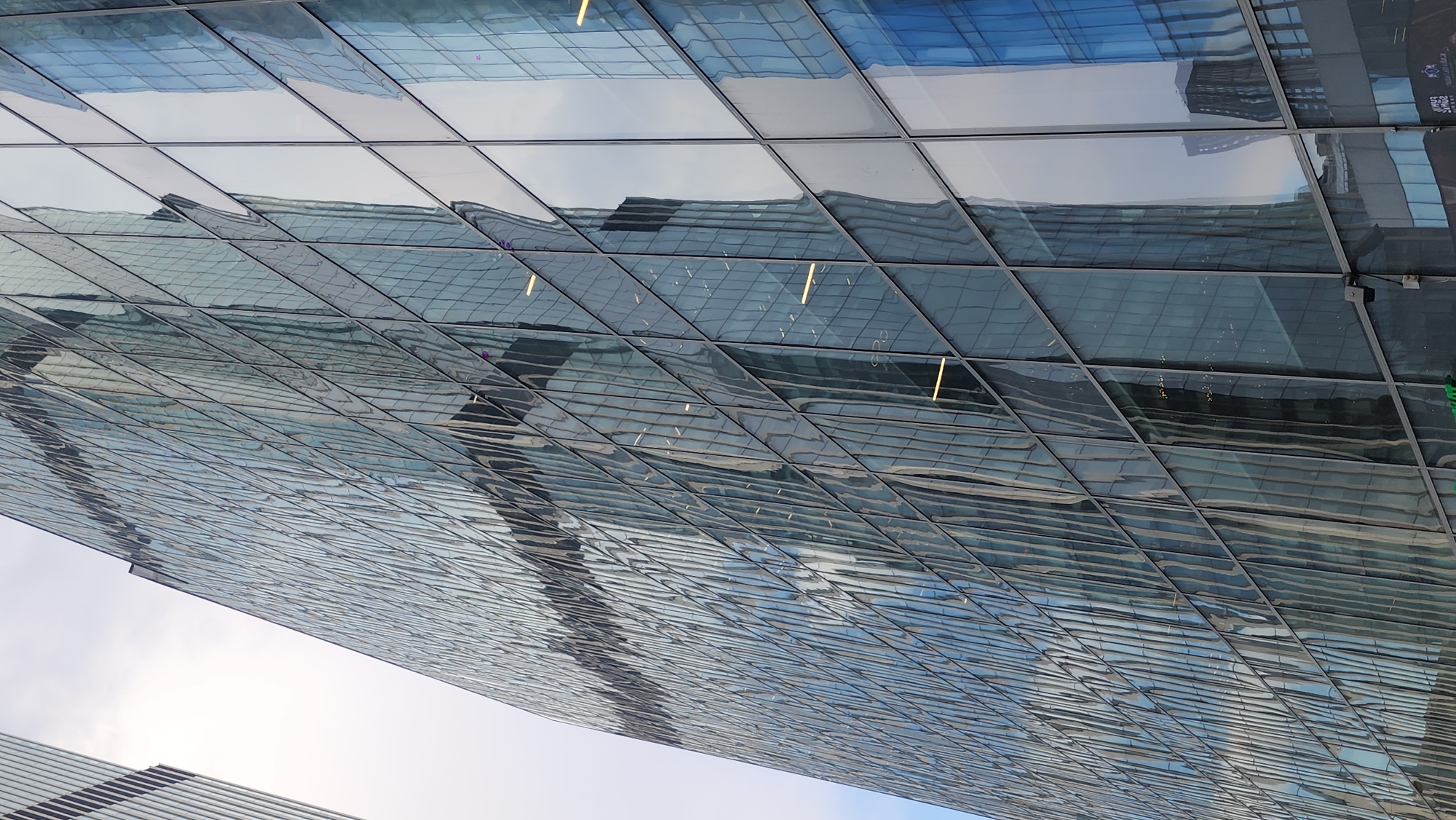 The image shows a portion of a modern glass-clad building, likely in the Moscow International Business Center (Moscow City) in Moscow, Russia. The architecture features a grid of reflective glass panels. The reflective surfaces display the sky and surrounding buildings. Some panels show interior lights on. A small portion of another building is visible at the bottom of the frame, also with similar glass panel construction. The sky is visible.