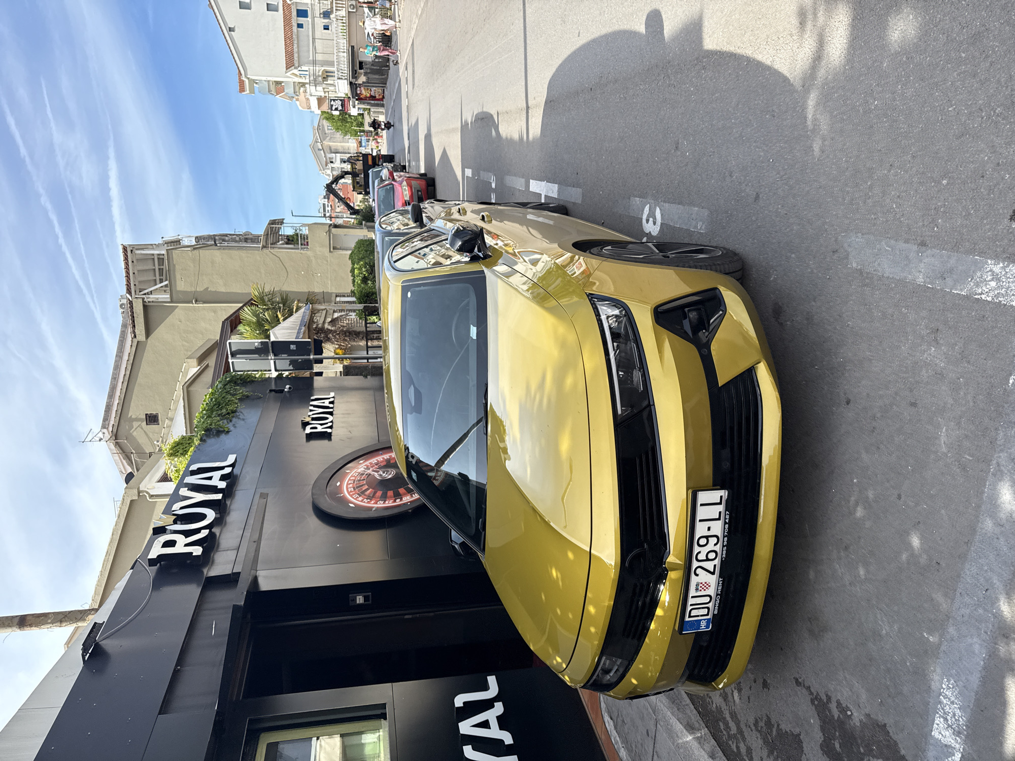 A vibrant yellow car is prominently parked on the side of a sunlit street in what appears to be an urban or coastal town, given the context of Budva, Montenegro. The car faces slightly left, showcasing its front grille and headlights. Its Croatian license plate, "DU 269-LL", is visible, along with text "BINGO RENT" and a Croatian phone number (+385 98 705 457).
On the left, directly beside the parked car, is a striking black building with the word "ROYAL" displayed in large white letters. A decorative, oversized roulette wheel, typical of a casino or gaming establishment, is affixed to its facade. Further down the street, beyond the yellow car, other vehicles are parked, including a red car and a darker one, with what looks like a recovery vehicle or vehicle with a crane arm positioned between them.
The street itself is paved, with white line markings indicating parking spaces, one of which clearly shows the number "3". Long shadows stretch across the asphalt, suggesting bright sunshine during late morning or early afternoon. In the background, several multi-story buildings line the street, with some having balconies and air conditioning units. A few pedestrians can be seen walking on the sidewalk. The sky above is a clear, bright blue, crisscrossed by several white contrails from aircraft. The overall scene depicts a typical sunny day in a busy town.