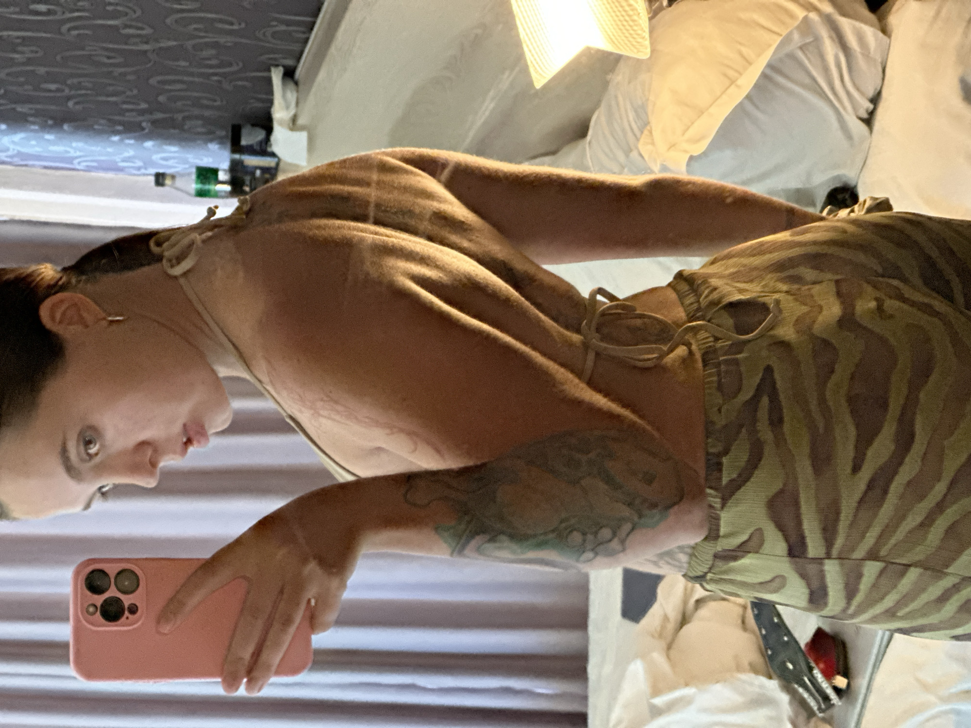 A woman is posing for a selfie in a hotel room. She is wearing a light-colored bikini top and patterned pajama pants. Her left arm is visible, and she has a tattoo on her upper arm. She is holding a pink smartphone in her left hand, which is capturing the image. The background shows a bed with white linens, a patterned wallpaper, and a lamp. The overall lighting suggests it is daytime or well-lit artificial lighting.