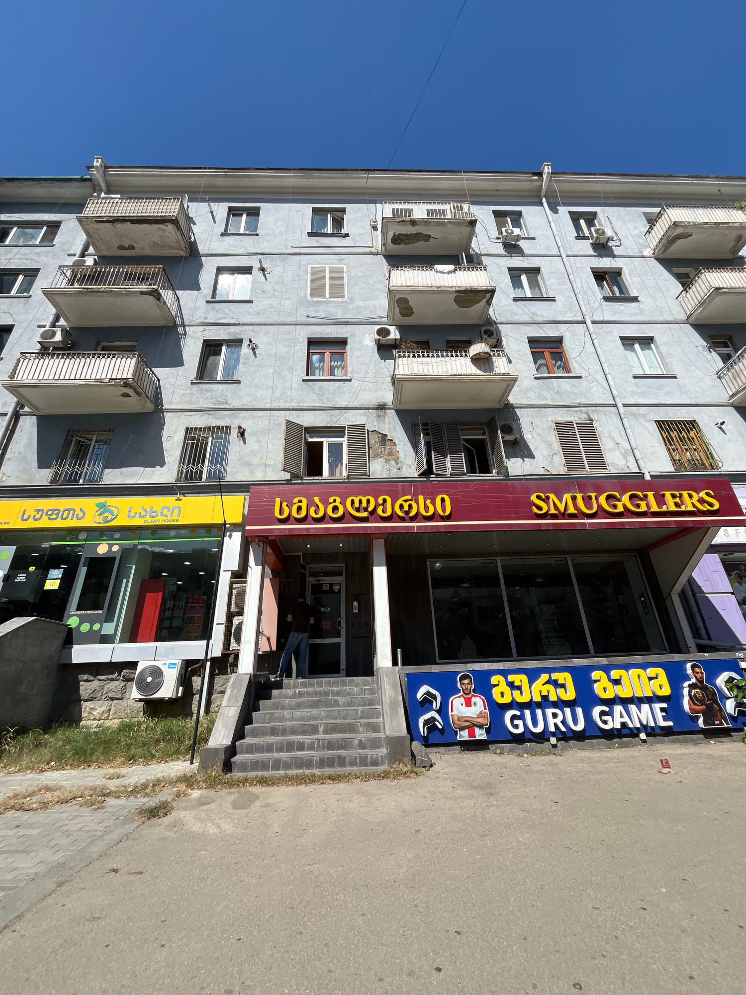 The image shows a street view in Tbilisi, Georgia, focusing on the facade of a multi-story apartment building with commercial spaces on the ground floor. The building is painted a light blue-grey color and features multiple balconies with rusted metal railings and deteriorating concrete. Air conditioning units are mounted on the exterior walls.
On the ground floor, there are two adjacent storefronts. The left storefront has a yellow sign with green and white text and logos, and large glass windows displaying items inside. Above its entrance, a red sign with yellow text reads "SMUGGLERS". To the right of the entrance, a blue sign with yellow and white text and images of athletes says "GURU GAME". A person wearing a dark shirt and blue jeans stands on the steps leading to the entrance of the "SMUGGLERS" store. The street in front of the buildings is paved with asphalt and concrete. The sky is clear and blue.