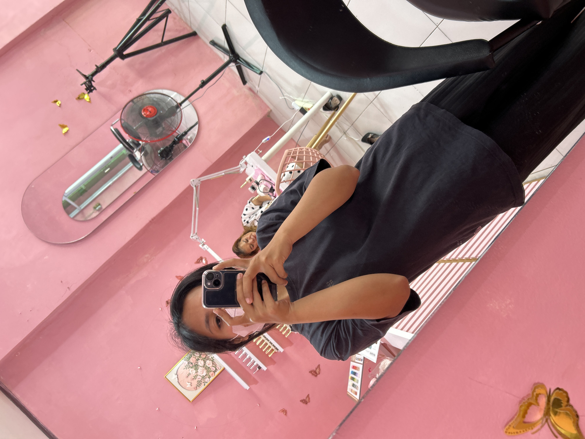 A young woman wearing a black t-shirt and a light pink face mask takes a selfie in a mirror. She is holding a black smartphone in her hands, with the camera facing her. In the reflection of the mirror, a second woman with dark hair is visible, lying down with her eyes closed. The room is decorated in shades of pink, with butterfly decals on the walls and shelves displaying various beauty products. A large oval mirror hangs on the wall, and a standing fan is positioned nearby. The scene appears to be a beauty salon or a similar establishment, potentially located in Depok, Indonesia.