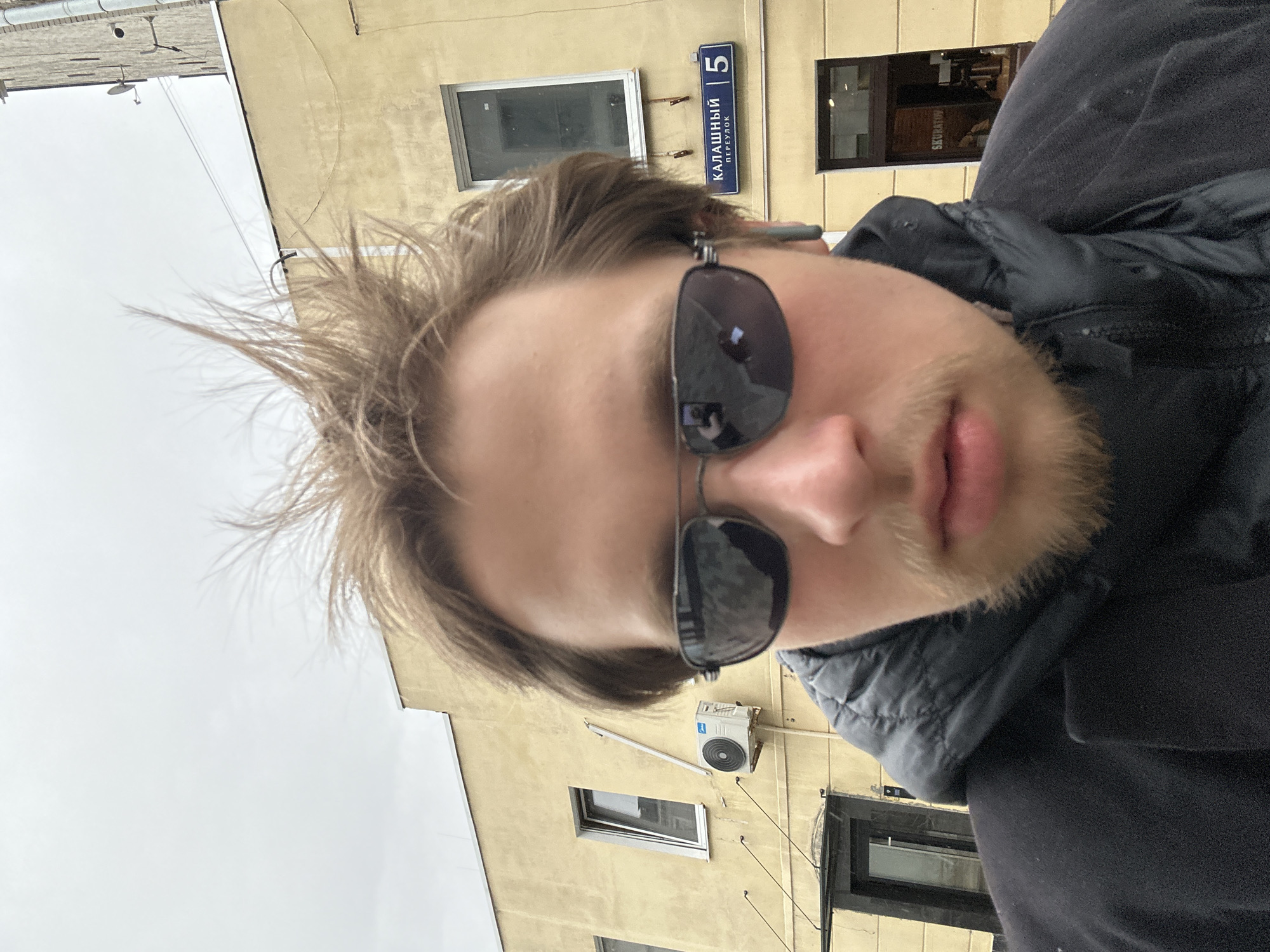 A close-up, tilted self-portrait of a person with blond hair and sunglasses. The person is wearing a dark-colored jacket. The background shows a yellow building with windows, an air conditioning unit, and a street sign that reads "Калашный переулок 5" (Kalashnyy Pereulok 5). The sky is overcast. The city and country are Unknown based on the information provided.