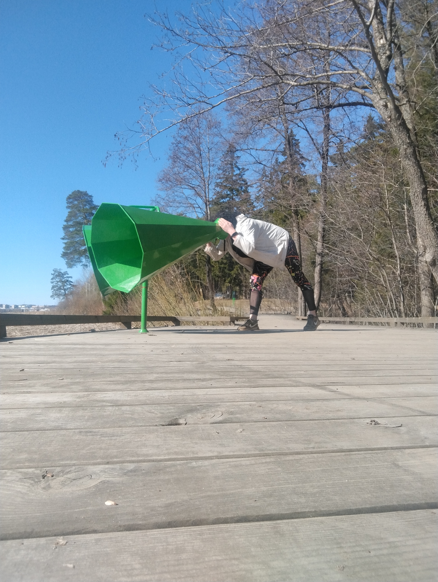 On a clear day in Votkinsk, Russia, a person in athletic leggings and a light-colored jacket bends over, placing their head inside one of two large, green, megaphone-shaped sound sculptures on a wooden boardwalk. The boardwalk is elevated and surrounded by a sparse forest of bare trees. In the distance, a cityscape is visible under the bright blue sky.