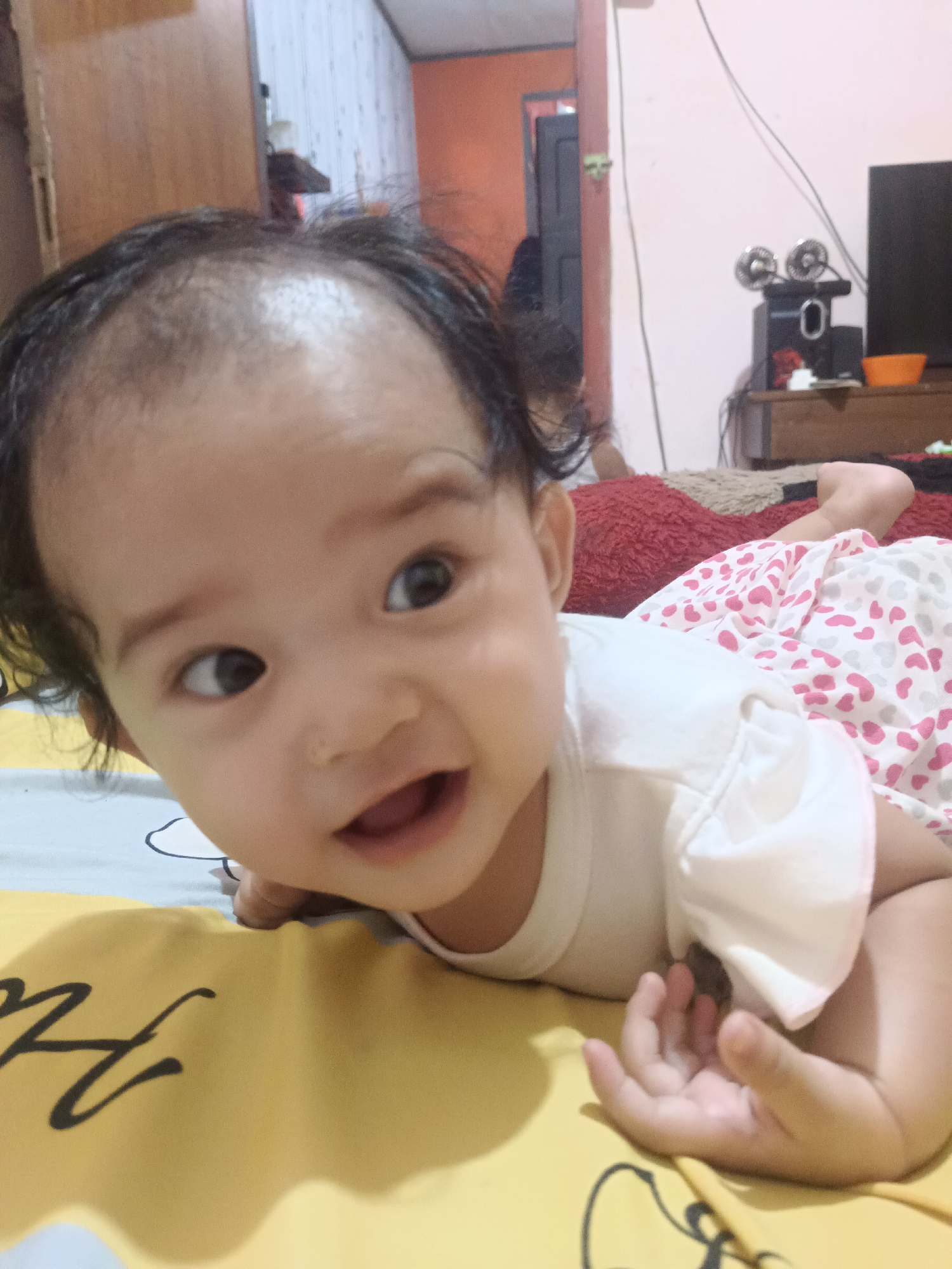 A young child, likely an infant, is lying on their stomach on a yellow and white patterned surface, possibly a bed or a play mat. The child has dark hair and is looking slightly to the left with wide, curious eyes. Their mouth is open, suggesting they are smiling or about to speak. They are wearing a white shirt with short sleeves and pink and white patterned shorts. The background shows a room with furniture, including a wooden dresser and a television on top of it. The overall impression is a candid, indoor photograph capturing a moment of playfulness. The location is specified as Kawua, Indonesia.