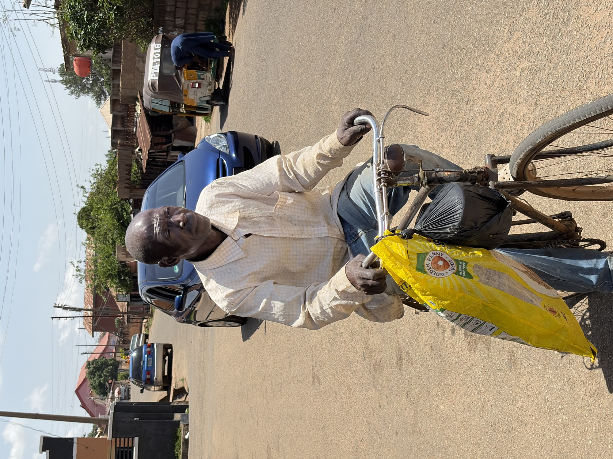 An older man rides or stands with his bicycle on a paved street in Jos, Nigeria, looking directly at the camera. He has a bald head, dark skin, and is wearing a light-colored, checkered long-sleeve shirt and blue jeans. His hands are on the bicycle's handlebars.
Attached to the bicycle is a large, bright yellow sack that prominently displays the text "PENNY SOYA" and "GOLDEN SUN" within a sun/flower logo, along with "TAI SOME A GOOD FOOD". A black plastic bag is also tied to the front of the bicycle. The bicycle itself appears to be a vintage model with a dark frame and a headlight on the front.
The scene depicts a sunny day with clear skies. In the background, parked cars (including two blue sedans) line the roadside. Further down the street, a green and yellow tricycle (keke napep), common in Nigeria, is visible with the alphanumeric "PTA-7018" on its rear window. A person in dark clothing is leaning into the back of the tricycle. Various residential and commercial buildings with reddish roofs, along with trees and overhead power lines, frame the street scene under bright daylight conditions. The road surface is light brown asphalt.