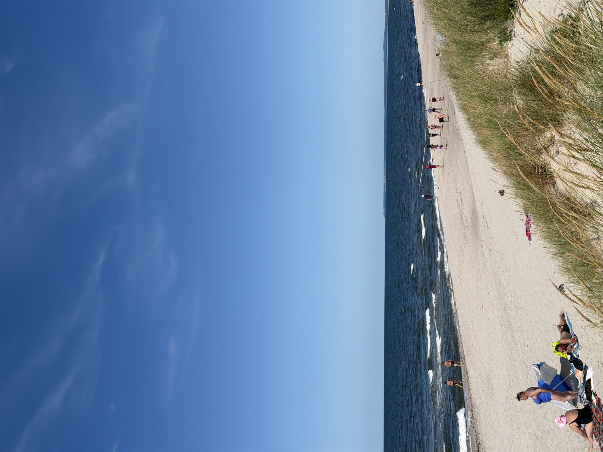 The image depicts a sunny day at a beach, likely in or near Baltiysk, Russia, given the presence of maritime elements. In the foreground, sand dunes with tall, dry grass are visible. Several people are scattered across the sandy beach. In the middle ground, a group of individuals are playing volleyball on the sand near the water's edge. The sea is a deep blue with moderate waves, and the sky is clear and bright blue with a few wispy clouds. In the distance, a shoreline is visible. A few individuals are in the water, and others are sunbathing or relaxing on towels. The overall atmosphere is one of leisure and outdoor recreation.
