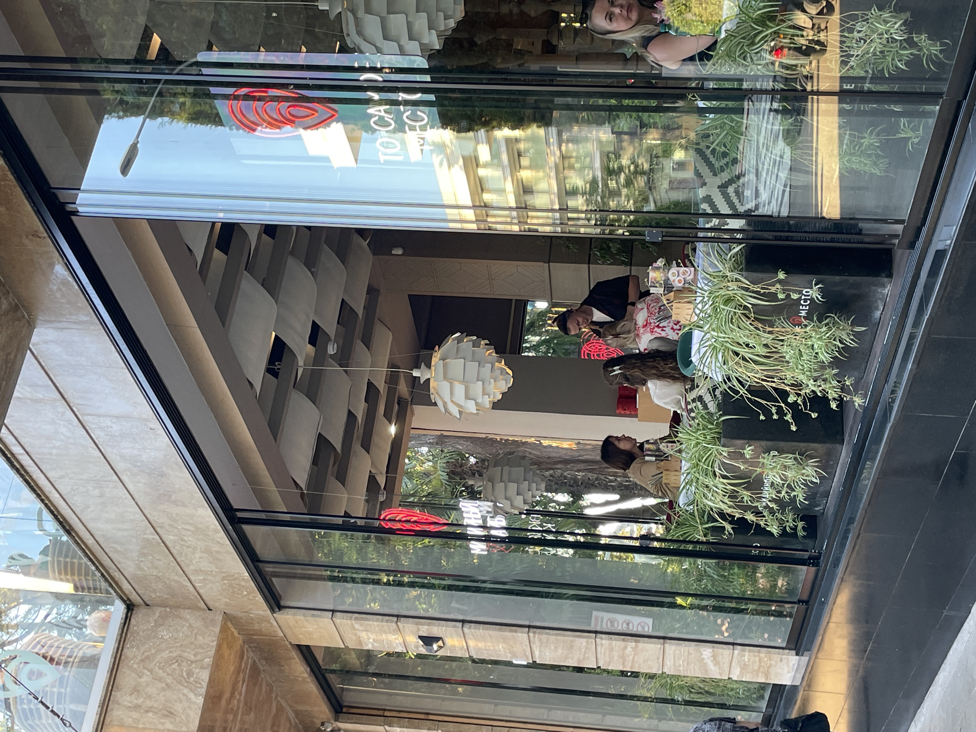 This media file captures a reflection of a street scene through the glass facade of a modern establishment. The primary subjects are people, observed through the glass, engaged in various activities. Inside, individuals appear to be seated at tables, suggesting a cafe or restaurant setting. One person, a woman with blonde hair, is visible looking towards the camera. Another individual, wearing a dark shirt and a beanie, is leaning over a table.
The scene is illuminated by natural light, indicating it is daytime. The reflections show the sky, trees, and buildings, hinting at an urban or semi-urban environment. The architecture of the building features large glass panels and a distinctive layered ceiling design visible in the reflection. Hanging light fixtures, one appearing as a large white geometric structure, are also visible. A prominent neon sign, glowing red in a stylized heart or flame shape, is reflected in the glass, along with other text that appears to be in Cyrillic script. One visible text reads "ТОТСА" and "MECTO".
The overall mood appears casual and relaxed, as people are seen enjoying themselves inside. The presence of potted plants adds a touch of greenery to the interior. The reflective nature of the glass creates a layered visual experience, blending the interior scene with the external environment.
