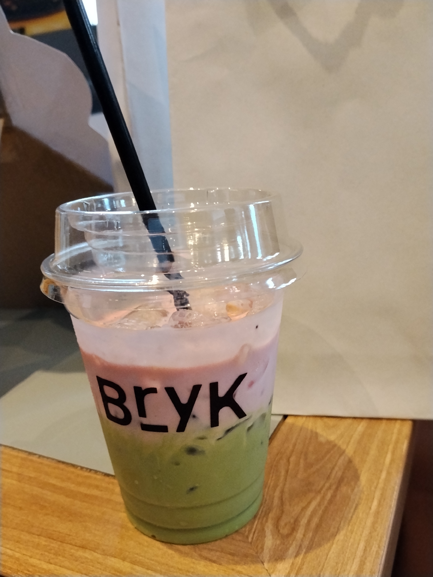 A close-up, eye-level shot shows a clear plastic cup filled with a layered drink, featuring a green bottom layer, a pink middle layer, and a light-colored foam top. A black straw extends from the cup. The cup has a clear plastic lid with embossed branding that reads "BRYK" in black capital letters. The drink sits on a wooden surface, with the background appearing to be a light-colored wall and a darker brown cardboard box. There are no people visible in the image, and the scene appears to be indoors, possibly a cafe or shop in Serang, Indonesia. The lighting is soft and natural, creating gentle shadows on the wooden surface.