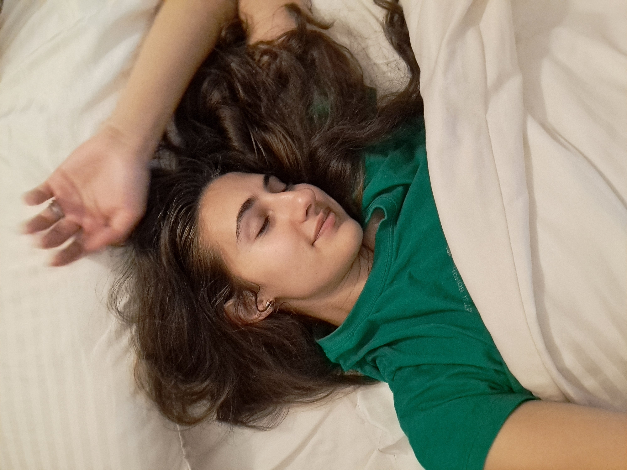 A person is lying in a bed covered in a white sheet. The person is wearing a green shirt. The person has long, brown hair that is spread out on the bed. The person's eyes are closed, and they appear to be sleeping. The person's left arm is raised above their head. The bed has a white, wrinkled sheet. The location is unspecified, and there is no information available that suggests this image was taken in the city of Negovan, Bulgaria.