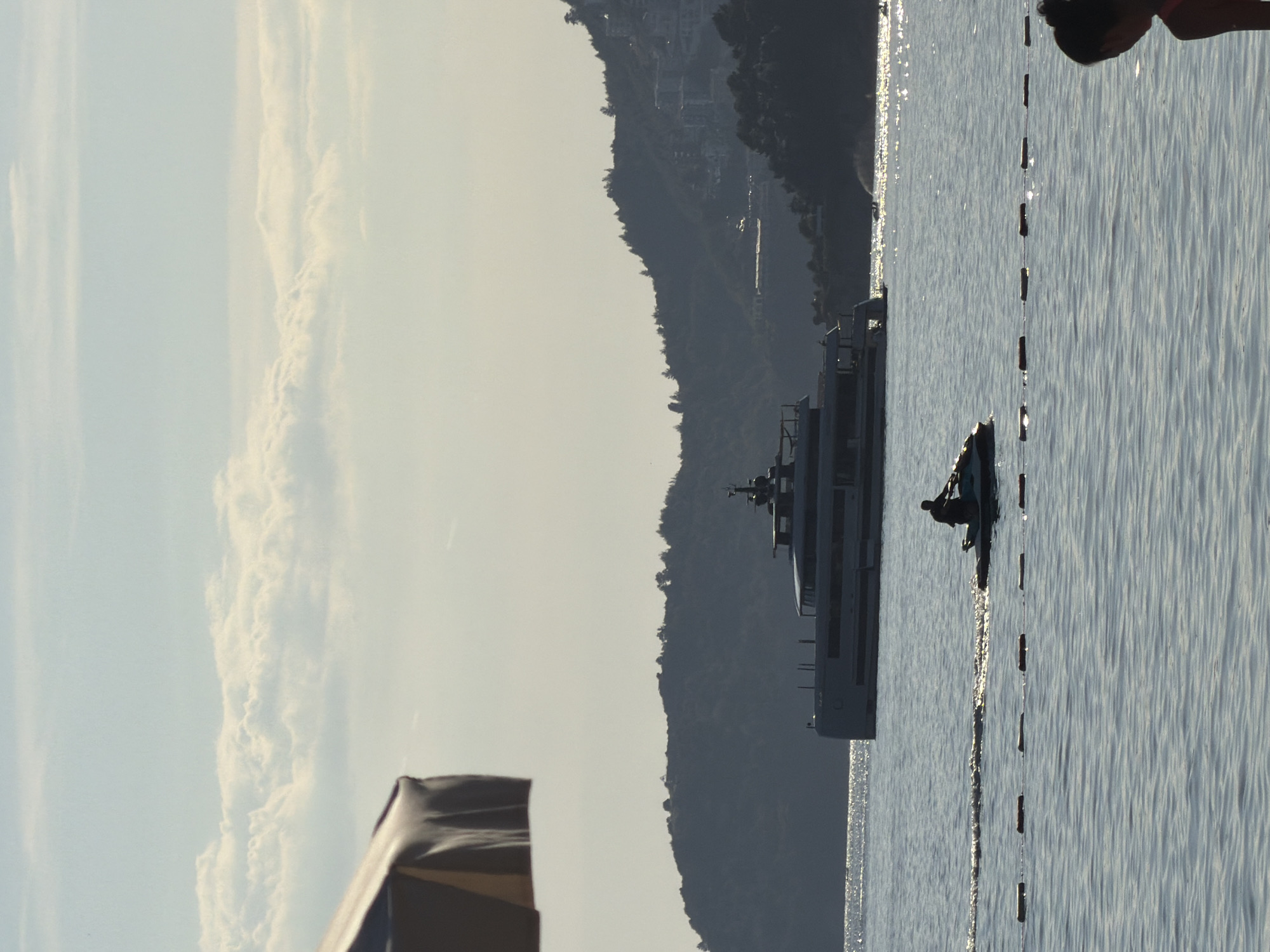 A landscape image shows a body of water, a mountain range, and a partially visible structure in the foreground. The water is calm with slight wave patterns and reflections of light. A line of buoys runs vertically through the water, creating a barrier. To the left of the water is a mountain range. A large ship is floating in the water near the mountain range. The sky is partially cloudy. The foreground contains a corner of an unidentified structure, partly obscured.
