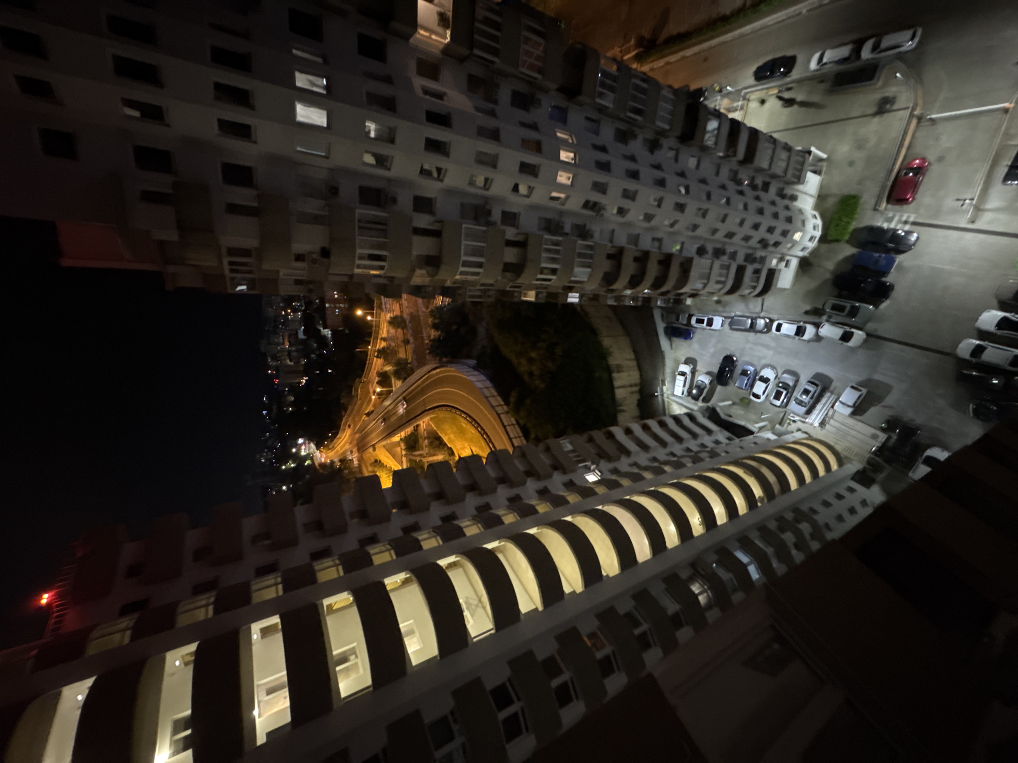 The image shows a high-angle night view of two residential apartment buildings in Sochi, Russia, with a highway visible in the background. The buildings are tall and have a curved, modern design with many balconies. Lights are on in some of the apartments, illuminating the interiors. A highway curves through the city, with cars visible on it. Parking lots are visible at the base of the buildings, with several cars parked. The sky is dark, indicating it is nighttime, and the city lights are visible in the distance. The overall scene depicts a well-lit urban environment at night.