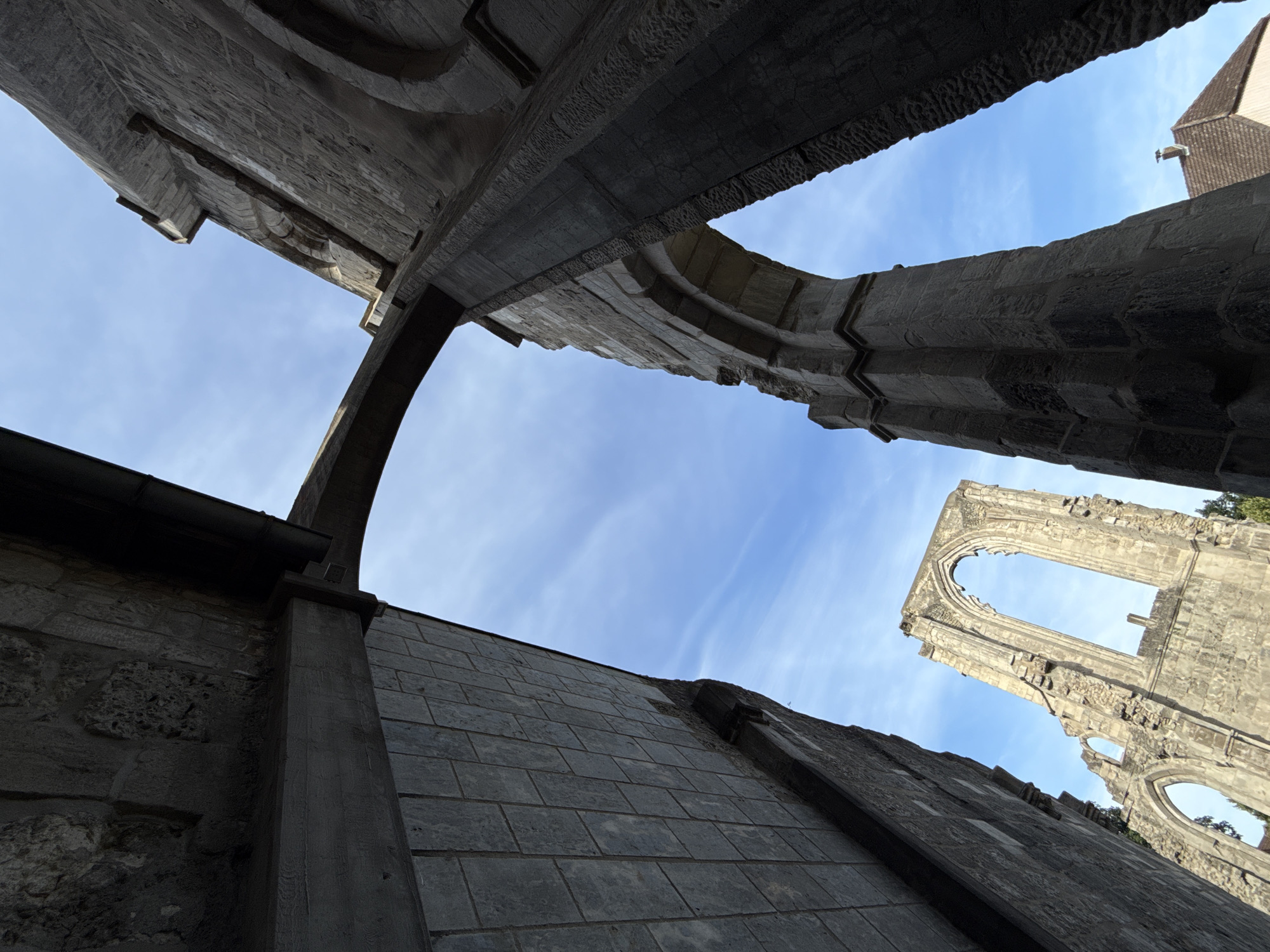 An extreme low-angle, wide-shot photograph captures the imposing stone ruins of Walkenried Abbey against a bright, clear blue sky with wispy white clouds. The perspective emphasizes the grand scale and architectural detail of the ancient structure, with massive stone arches and weathered walls dominating the frame. In the foreground, a dark, rough-hewn stone column on the right and a section of a finely tiled wall on the left create a dramatic visual contrast. In the mid-ground, a large arched window, characteristic of Gothic architecture, stands as a testament to the abbey's former glory. In the background, the roof of a smaller, more modern building with a reddish-brown tiled roof and a chimney peeks out from behind the ruins on the right. The image conveys a sense of historical grandeur and timelessness, highlighting the enduring presence of the Walkenried Abbey ruins in its German setting. There are no people or animals visible in the image.