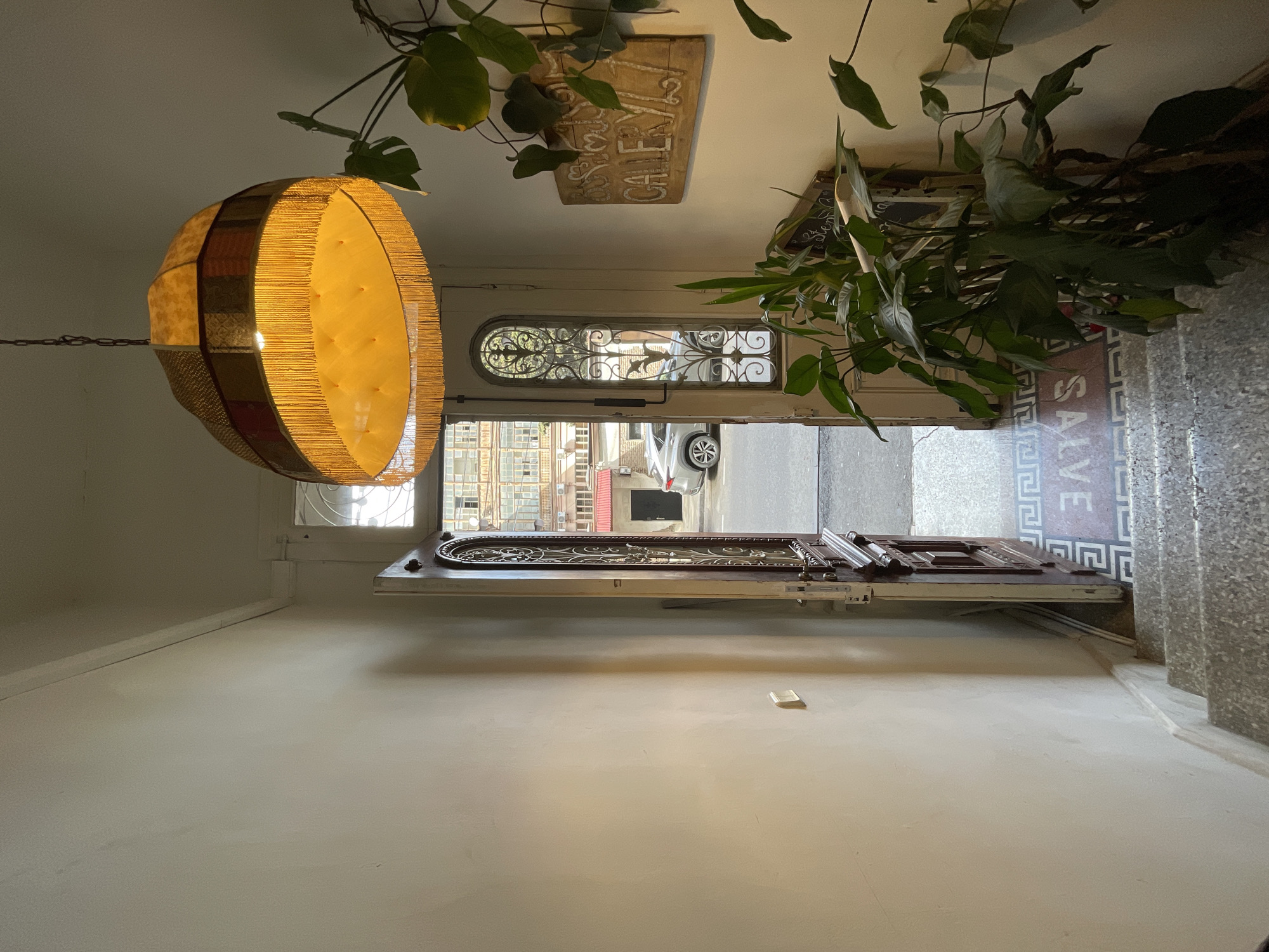 The image depicts an interior view looking outwards through an open doorway. The doorway leads to a street scene, revealing a building across the street with visible windows and architectural details, plus a parked white vehicle. The foreground features a light-colored wall, a hanging lamp made of woven material, and indoor plants partially obscuring the upper portion of the doorway. The flooring transitions from an interior tile pattern to exterior stone steps. A wooden sign hangs near the doorway, partially obscured by the plants. Based on the architectural style, the location is likely in Tbilisi, Georgia. No people are visible.