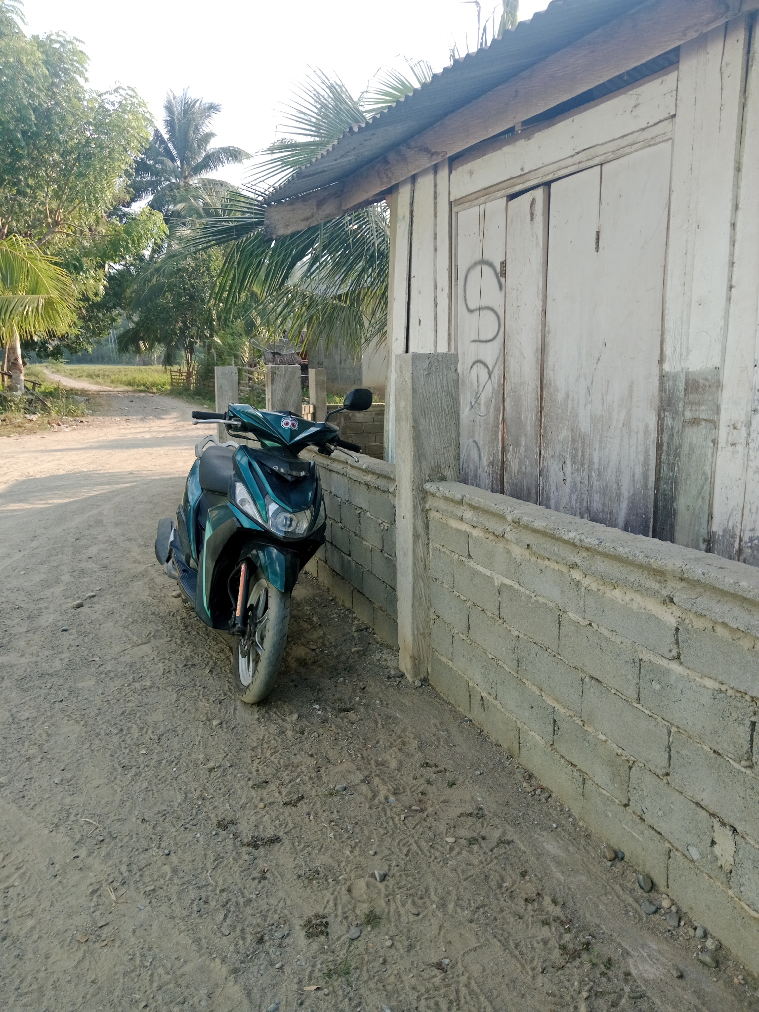 A teal and black scooter is parked on a dirt road in front of a simple wooden building with a corrugated metal roof. The building has white vertical siding and what appears to be a sliding door with a graffiti tag. A low concrete block wall runs along the front of the building and partially obstructs the view of the road. Trees, including palm trees, are visible in the background, suggesting a tropical or subtropical environment. The lighting indicates it is daytime, likely morning or late afternoon due to the long shadows. The scene appears to be in a rural or semi-rural setting, possibly in Lakea Satu, Indonesia, based on the provided context. There are no people visible in the image.